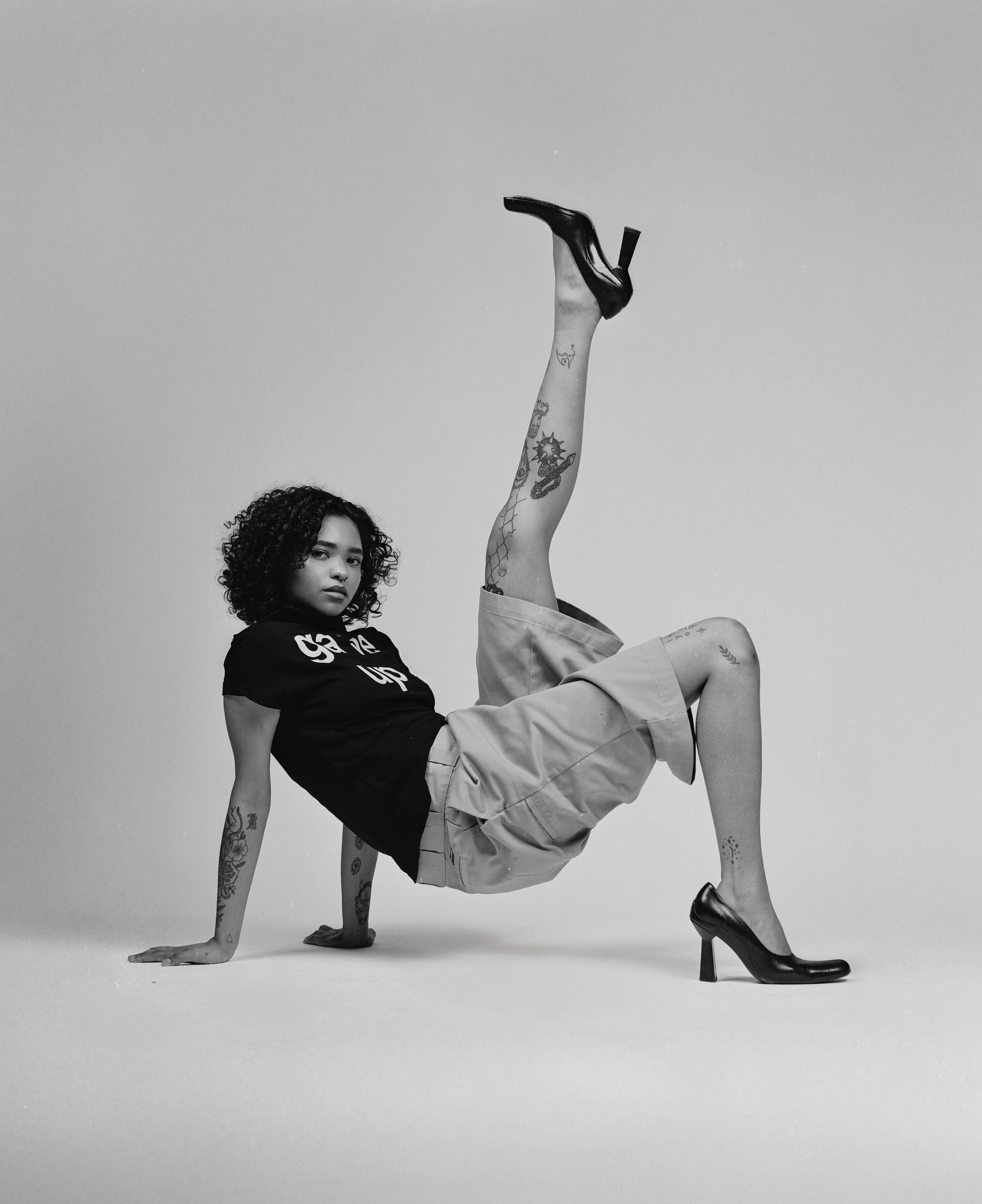 A young woman with curly hair and tattoos poses in a studio, wearing a black T-shirt with lettering, beige shorts, and high heels, with one leg raised vertically and her hands on the ground.