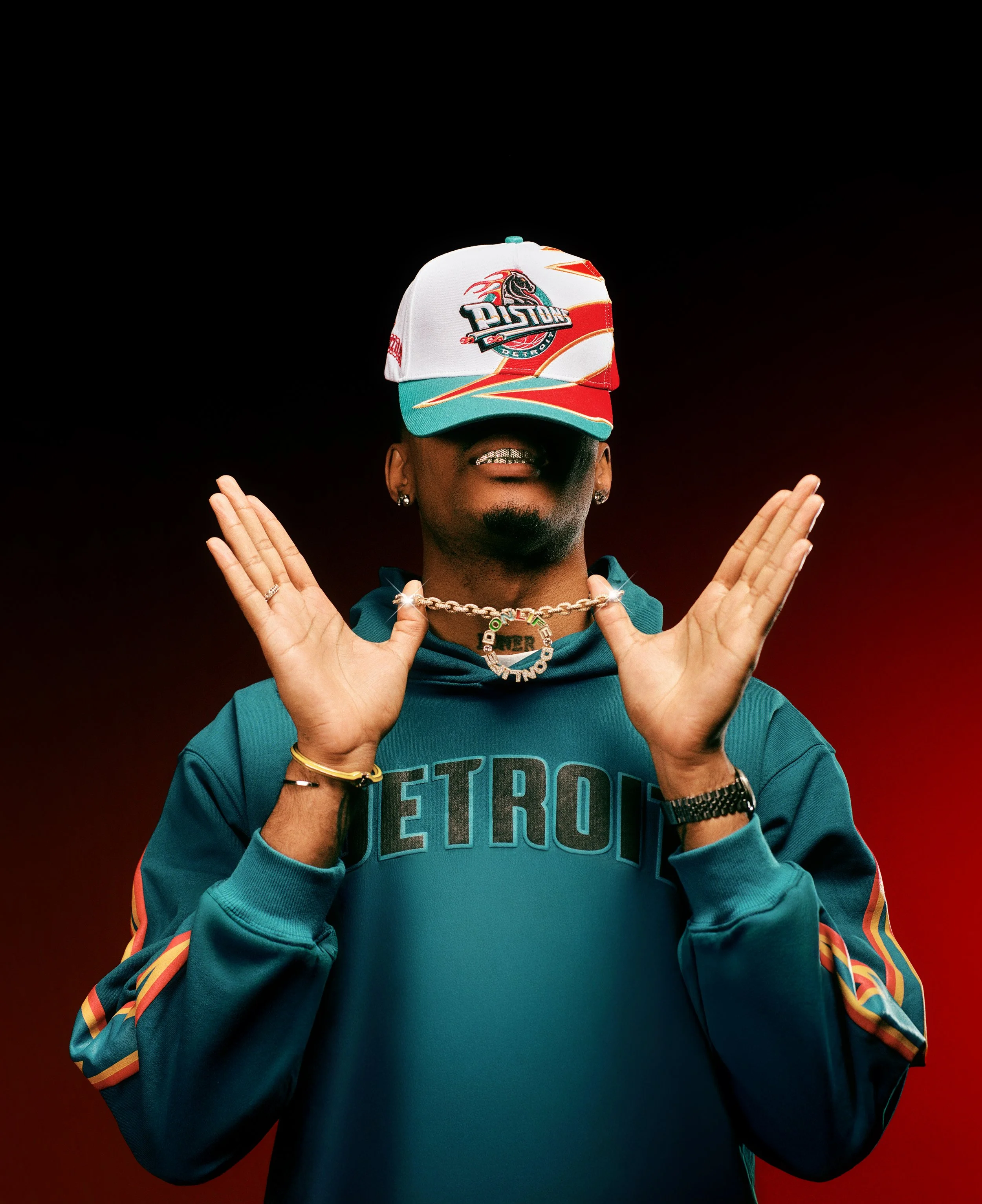A man wearing a Detroit Pistons baseball cap, gold grill, jewelry, and a teal Detroit hoodie, with a dark background.