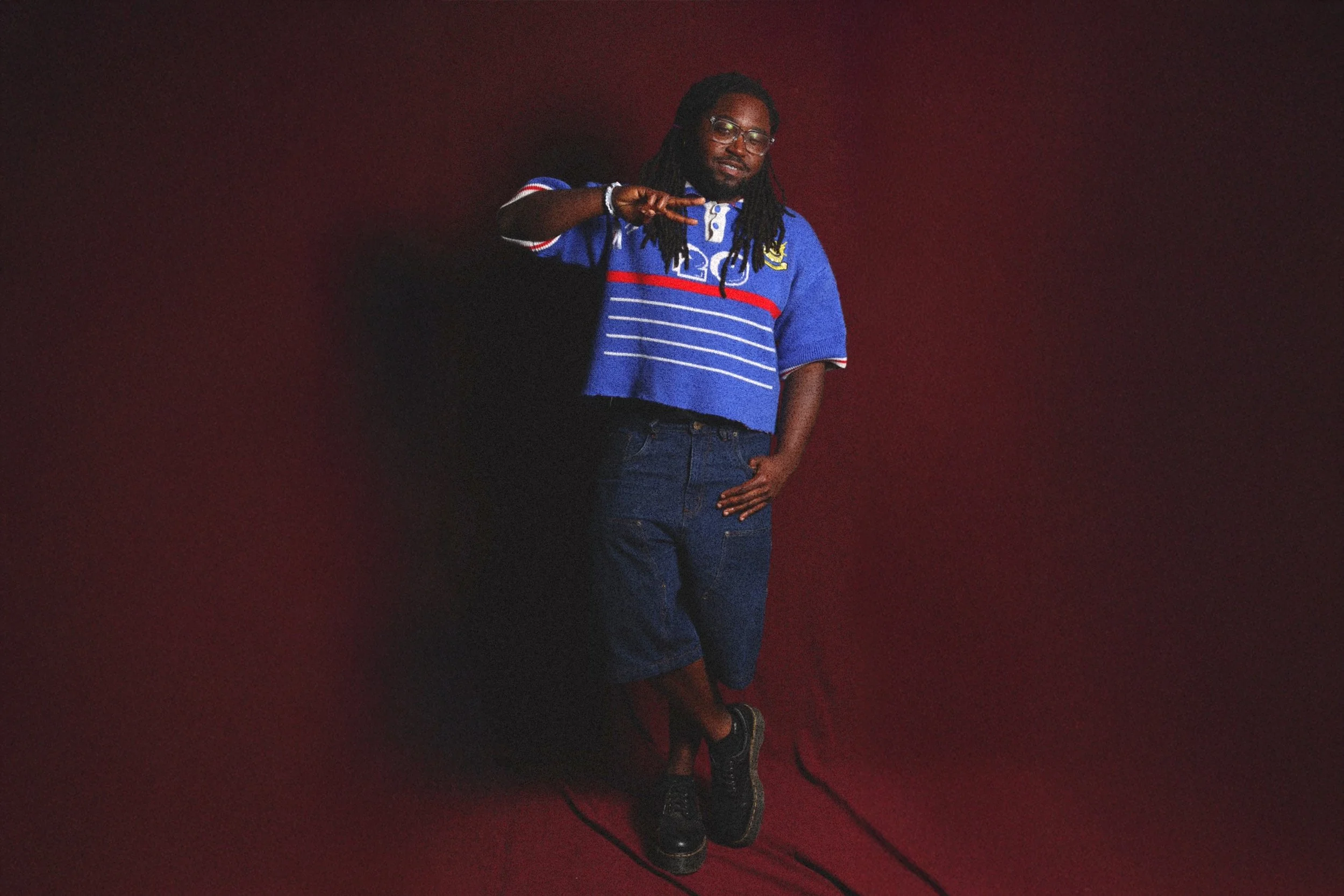 A man with long dreadlocks, wearing glasses, a blue polo shirt with red and white stripes, blue jeans, and black shoes, standing against a red background, making a gesture with his right hand.