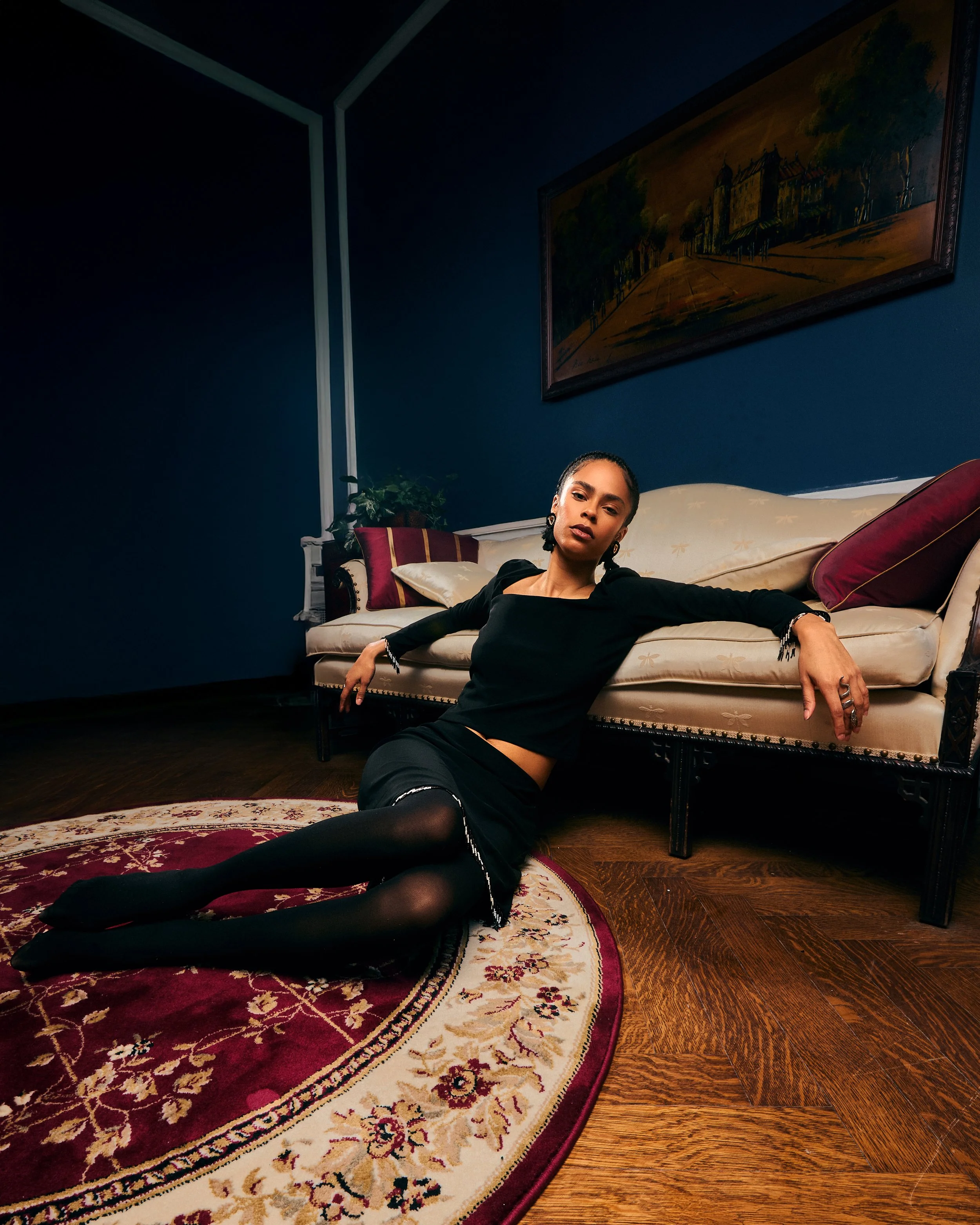A woman dressed in a black outfit with sheer black tights and a black skirt with white trim, sitting and lying on a patterned area rug in a dimly lit room with navy blue walls, a cream-colored vintage sofa with red and purple pillows, a painting hang