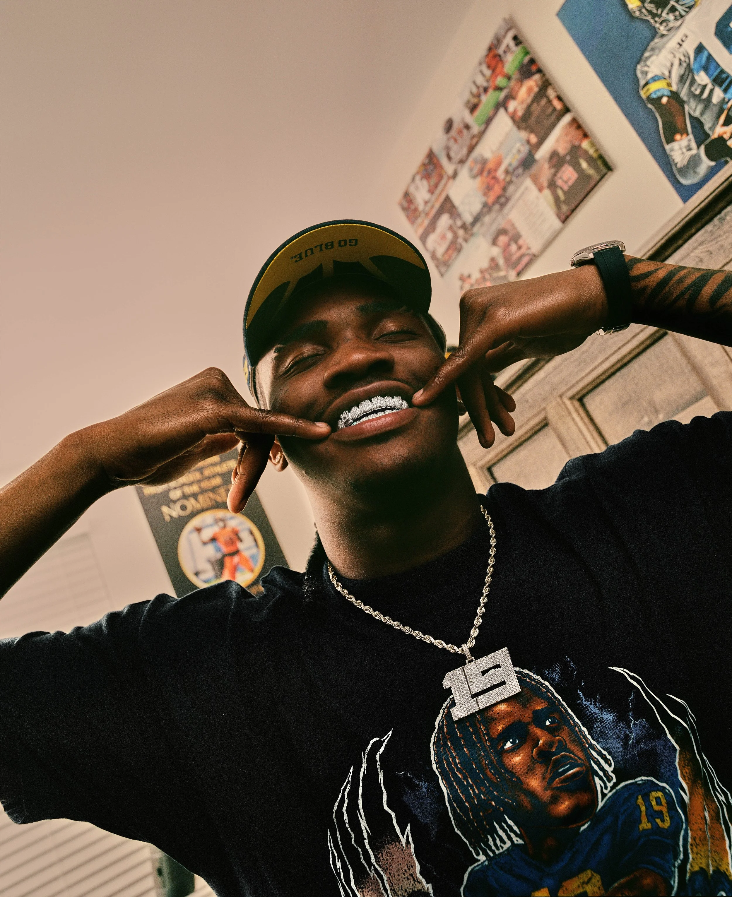 Young man wearing a yellow cap, smiling, showing teeth with a grill, holding his cheeks with fingers, wearing a black t-shirt with a graphic of a person in a football uniform and a chain with a large letter 'E' pendant, in a room with posters and pic