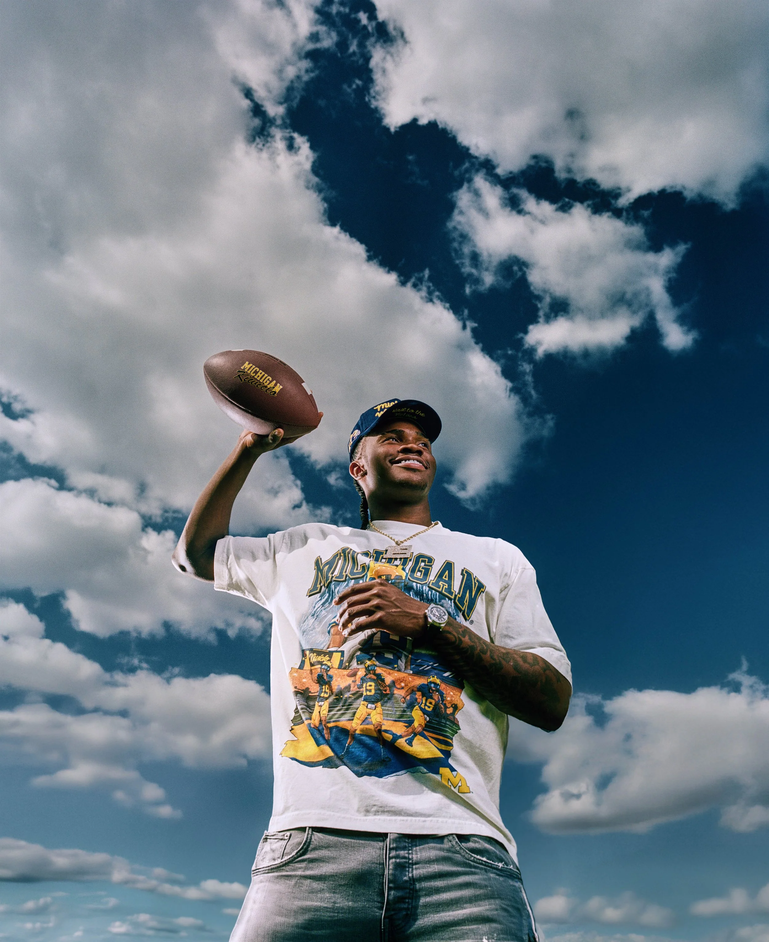 A young man holding a football while standing outdoors under a partly cloudy sky, wearing a Michigan football T-shirt and a baseball cap.