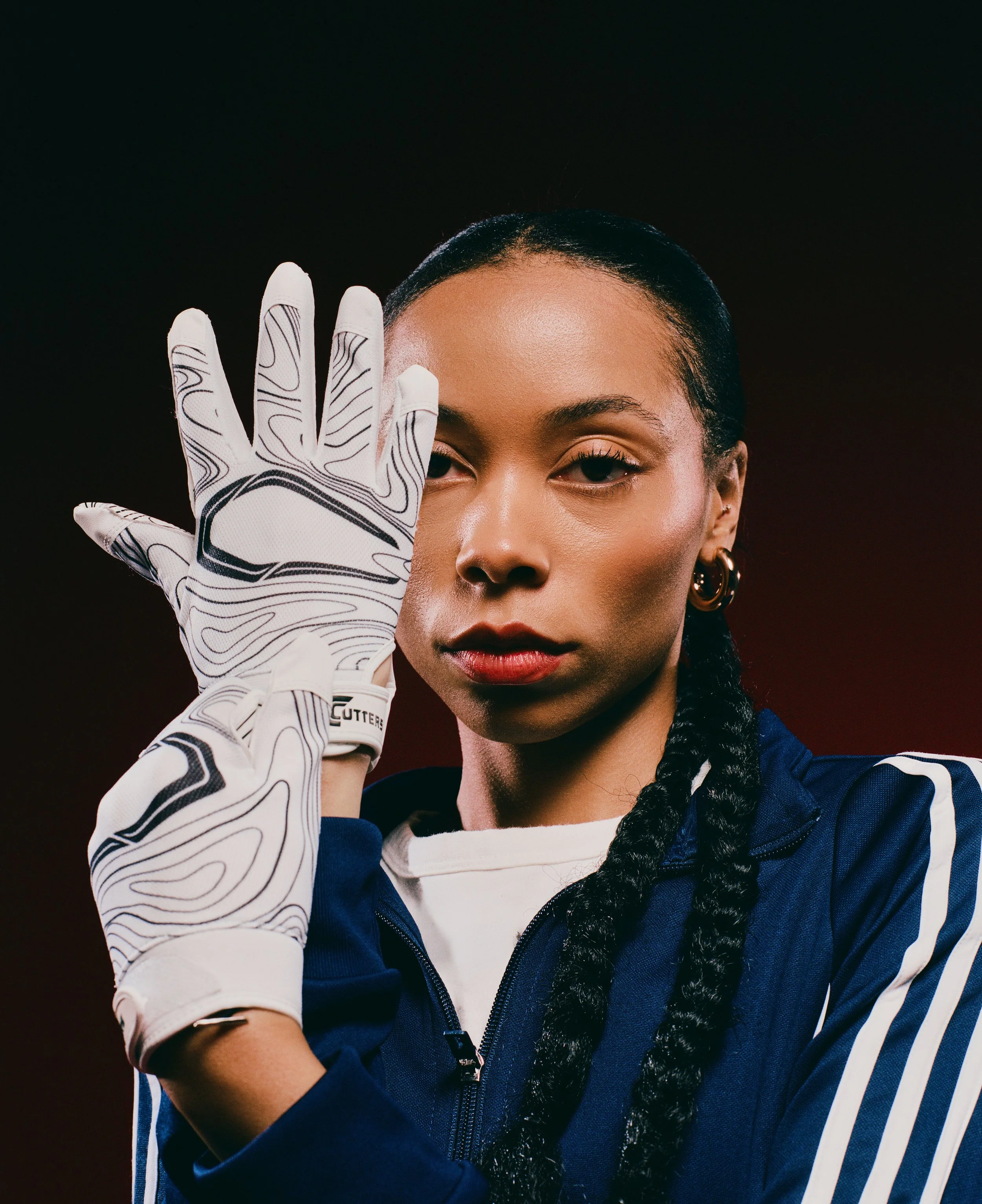 A woman in a blue sports jacket partially zipped, wearing white gloves with black abstract lines, holding her left hand up to her face with her fingers extended, against a dark background.