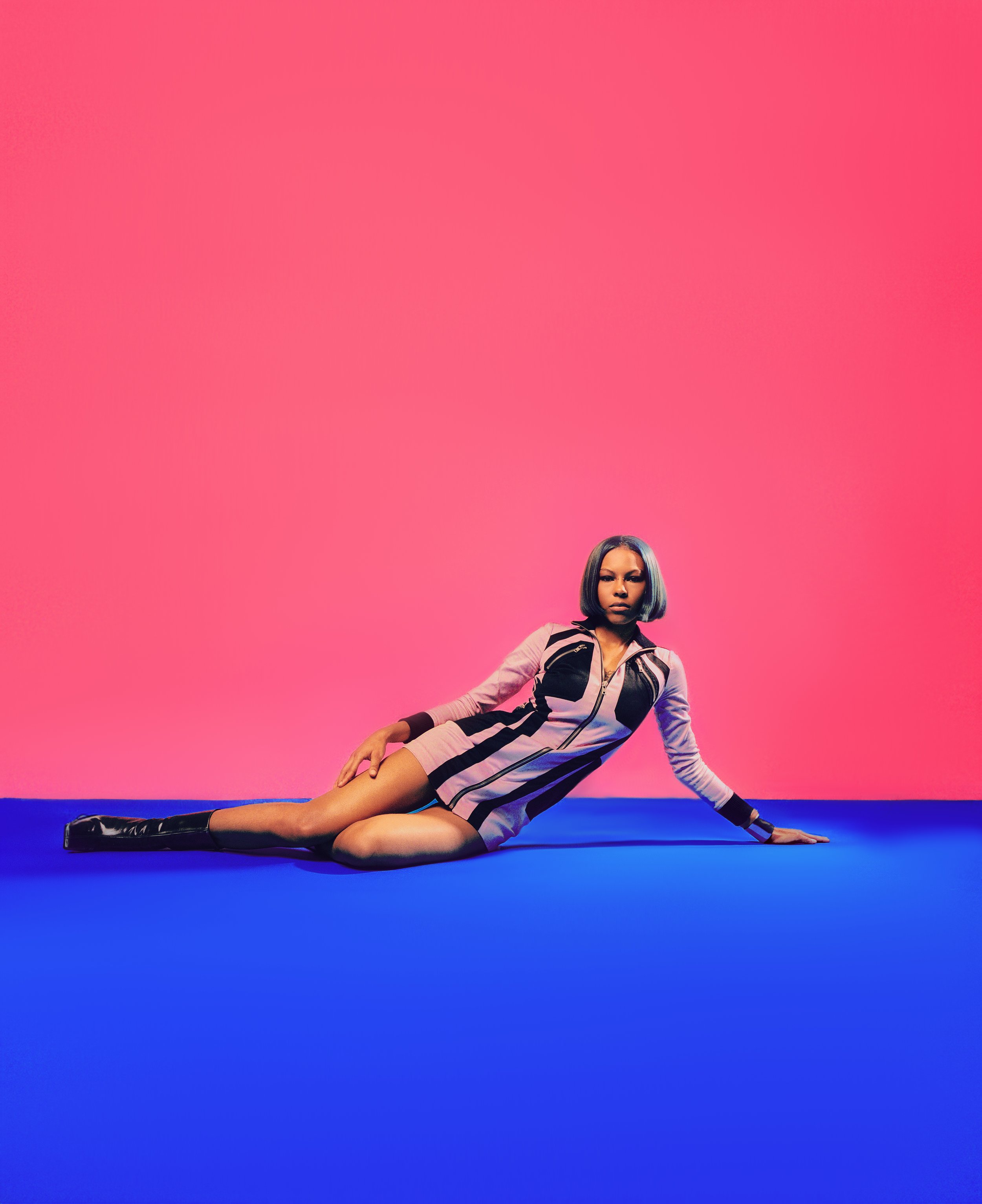 A woman with dark hair in a bob hairstyle, in a futuristic outfit, is lying on the floor against a pink and purple background.