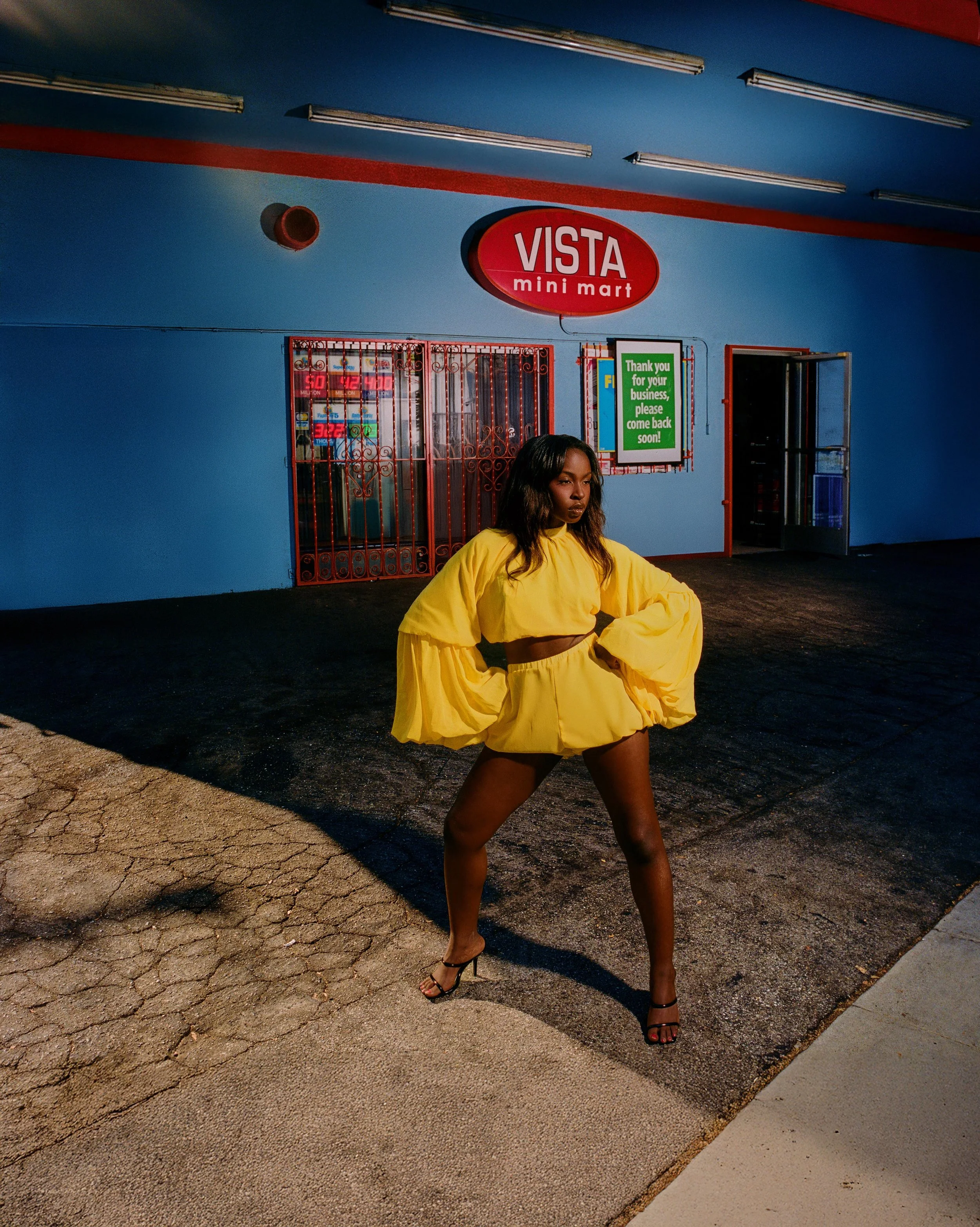A woman in a bright yellow outfit with black high heels standing outside a blue mini-mart called Vista Mini Mart, with a red sign and a green promotional sign on the wall.