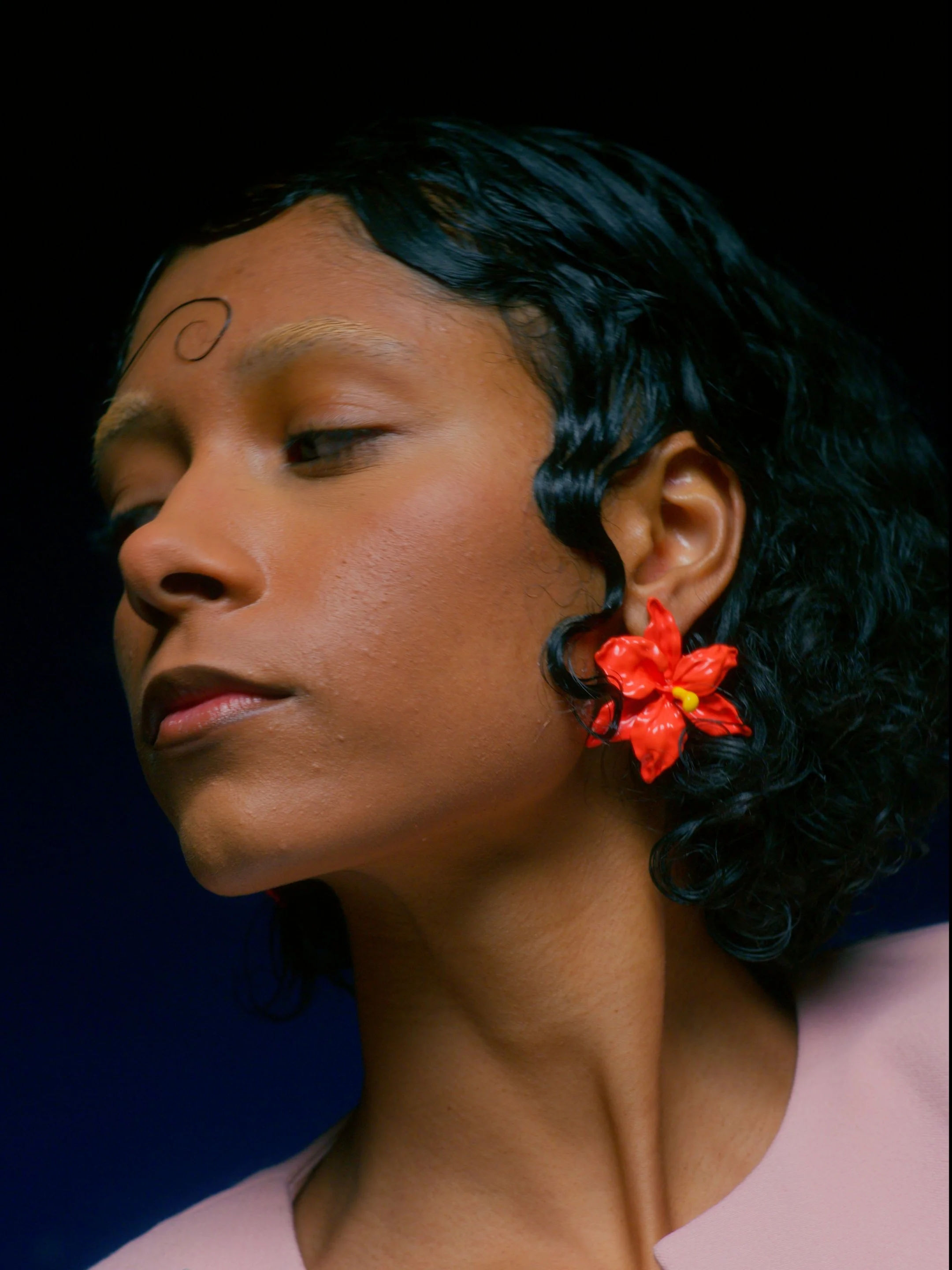 A woman with dark curly hair, wearing a red flower earring and a light pink top, with her eyes closed and head slightly tilted.