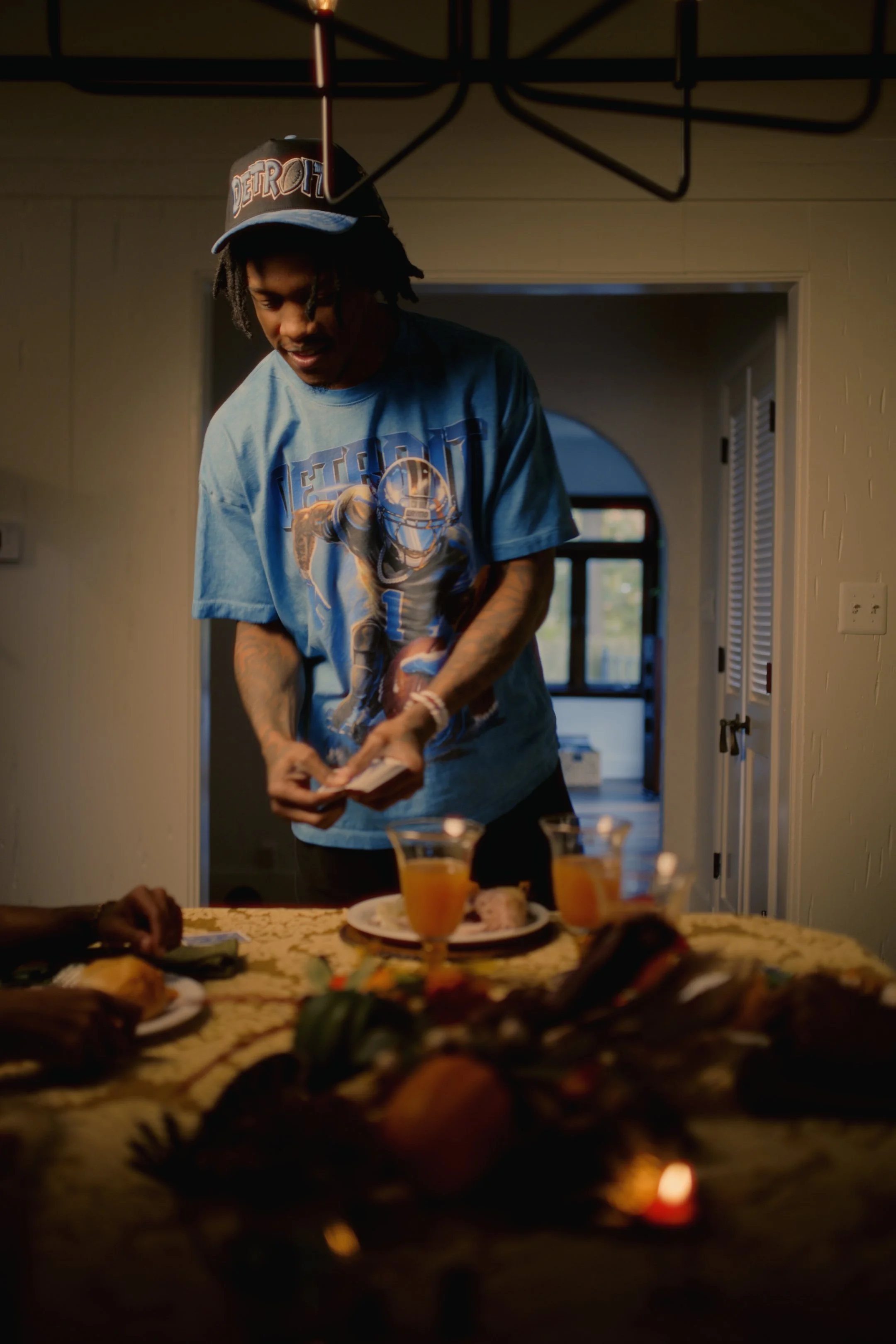 A young man wearing a Detroit Lions football shirt and a cap is preparing to lay money on a table set with food and drinks, with a plural arrangement of cooked food and two glasses of orange juice in the foreground, in a warm lit dining room.
