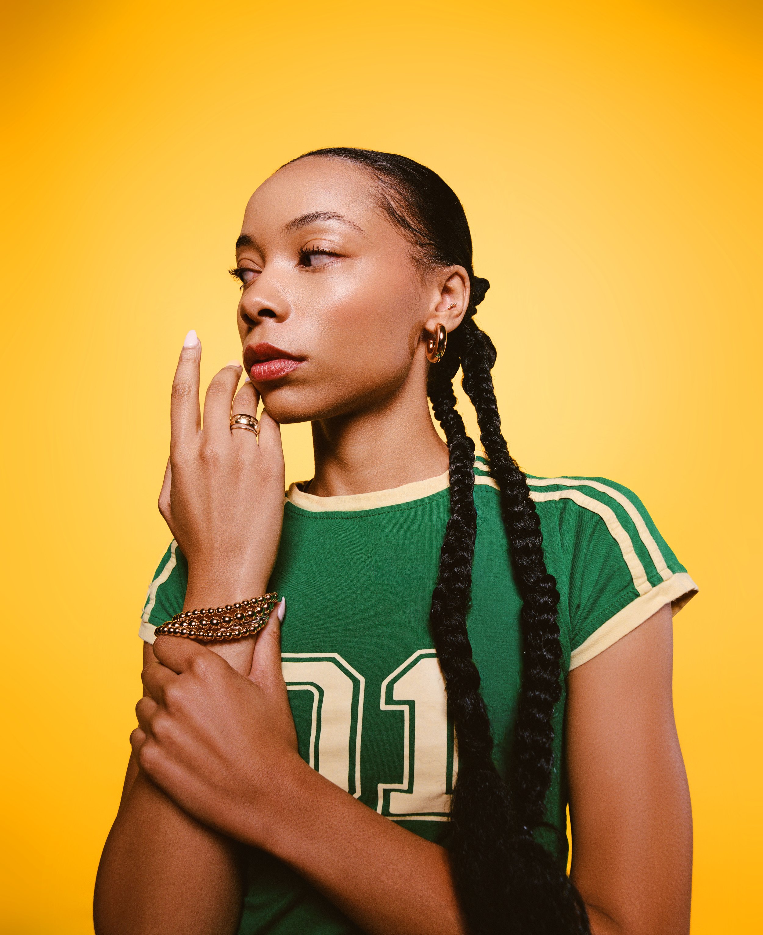 A woman with long braids stands against a yellow background, wearing a green sports jersey with yellow accents, gold jewelry, and a thoughtful expression.