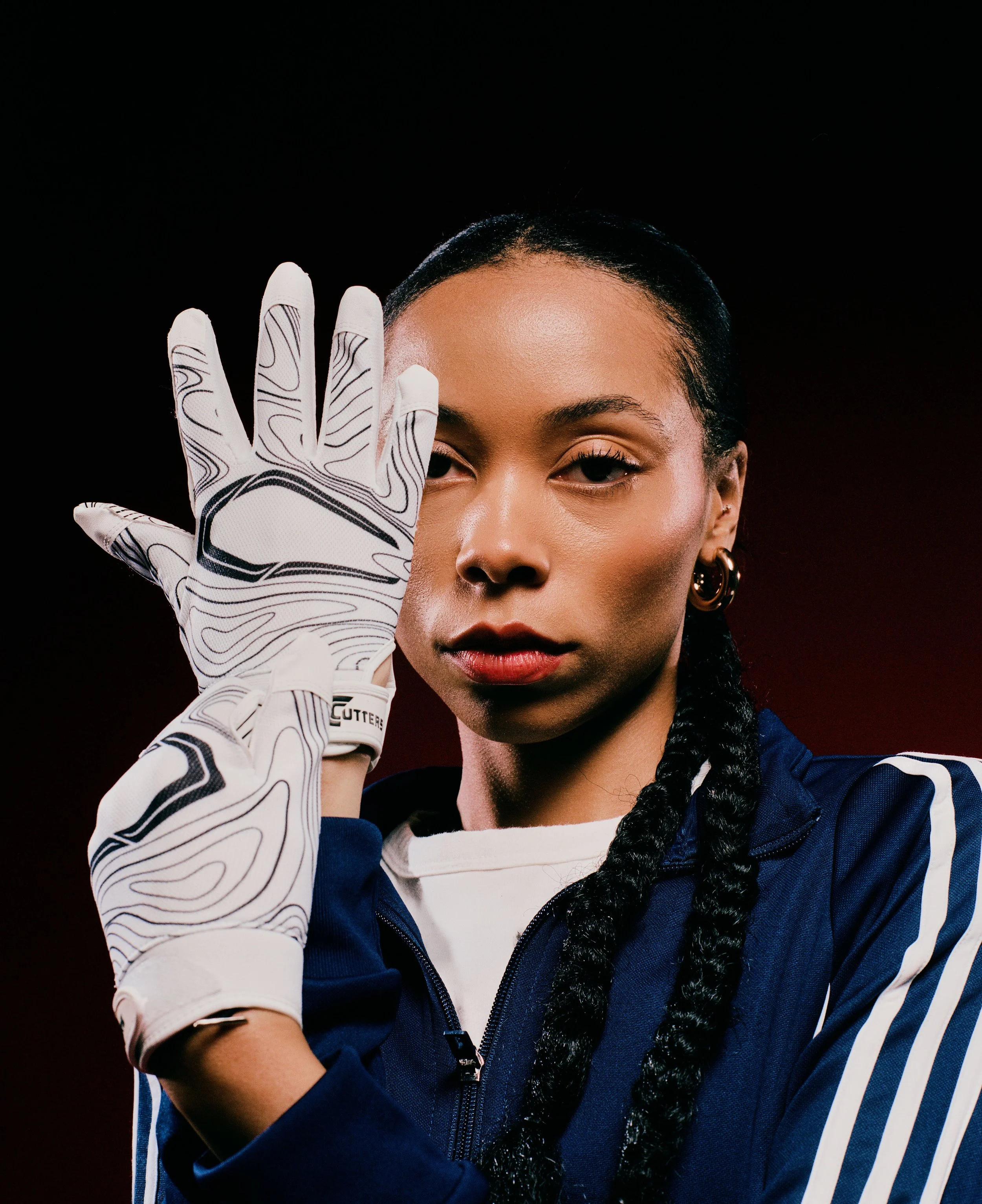 A woman with braided hair wearing a blue sports jacket and a white glove with black designs, posing with her hand near her face against a dark background.