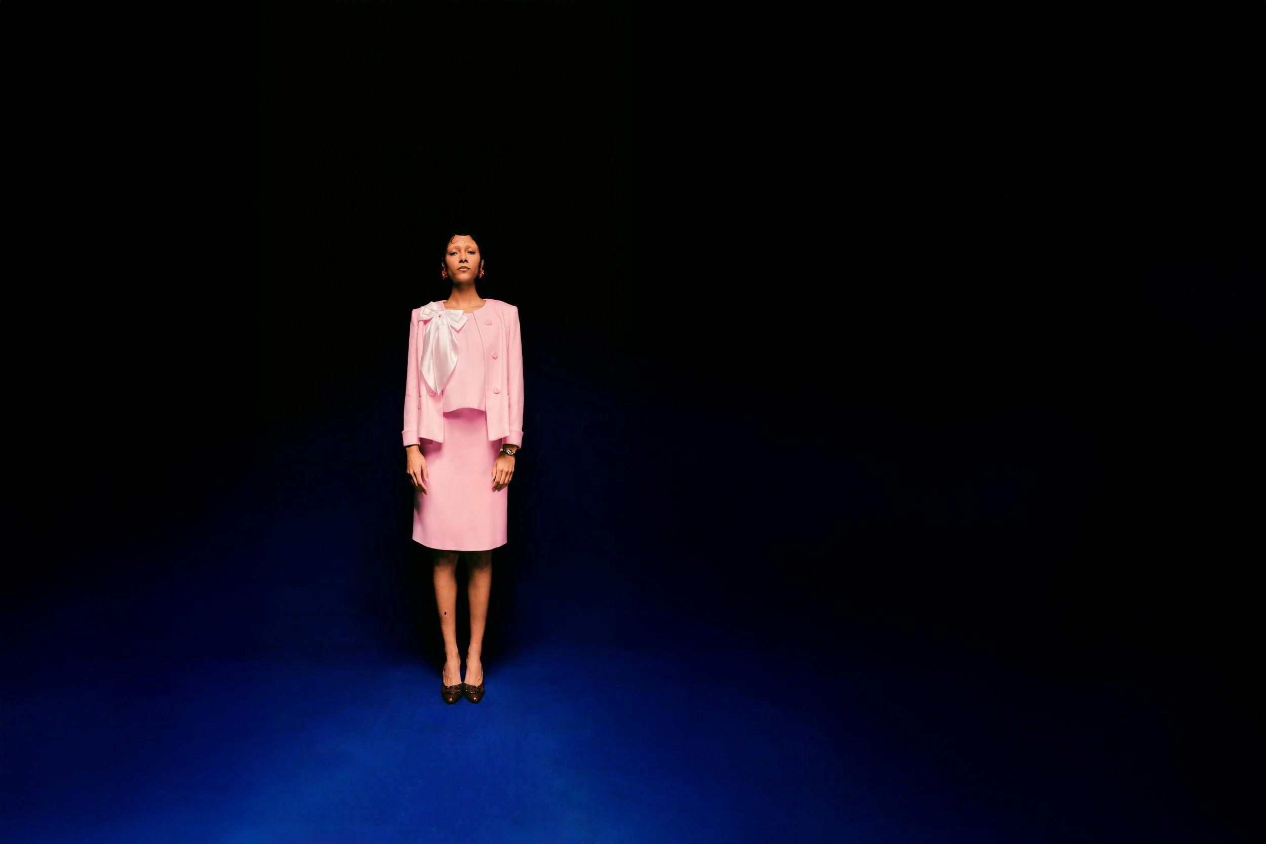 A woman wearing a pink dress and a pink blazer standing on a blue stage with a black background.