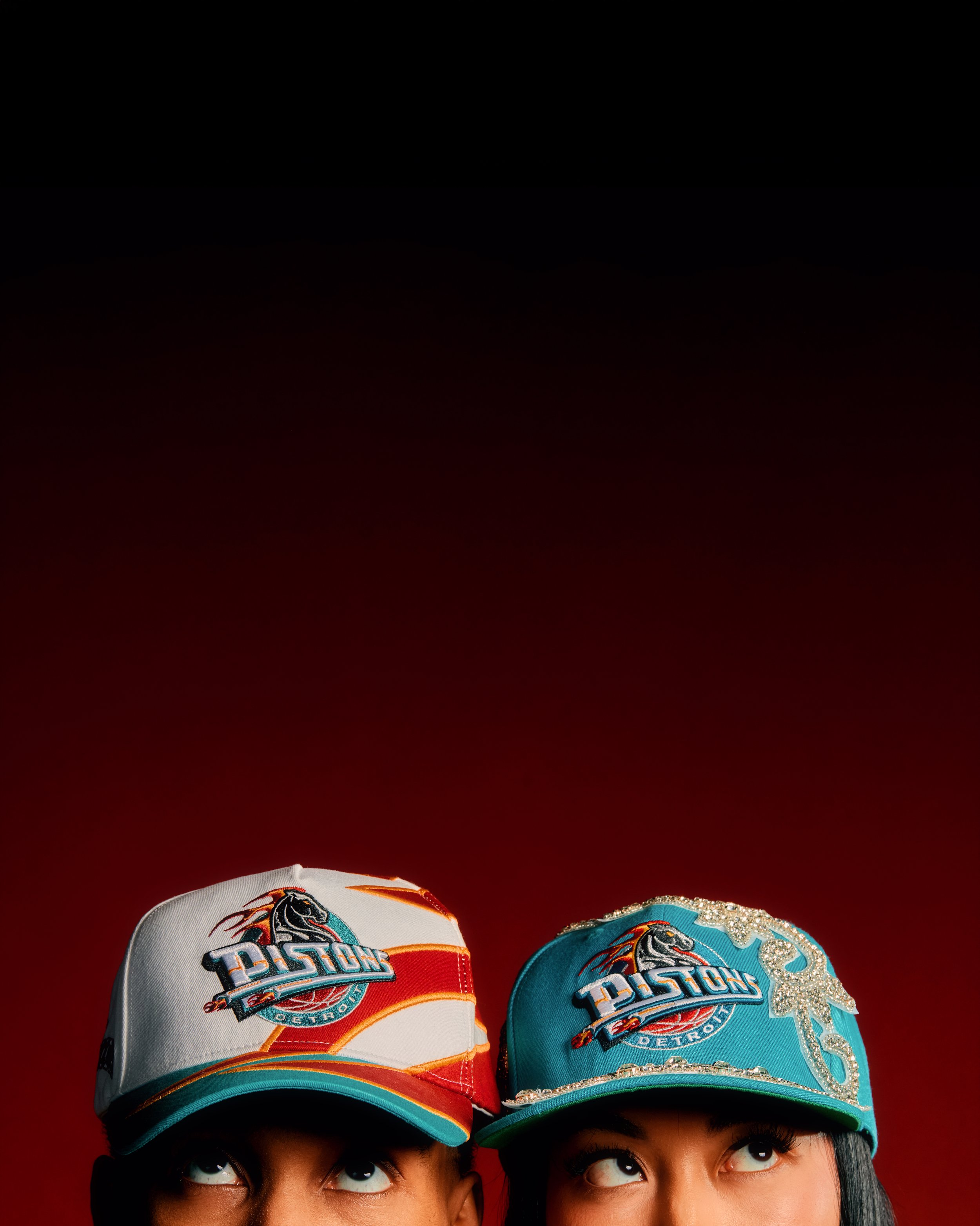 Two women are wearing Detroit Pistons basketball caps and looking upwards. One cap is white with a sequin design, and the other is teal with the Pistons logo.