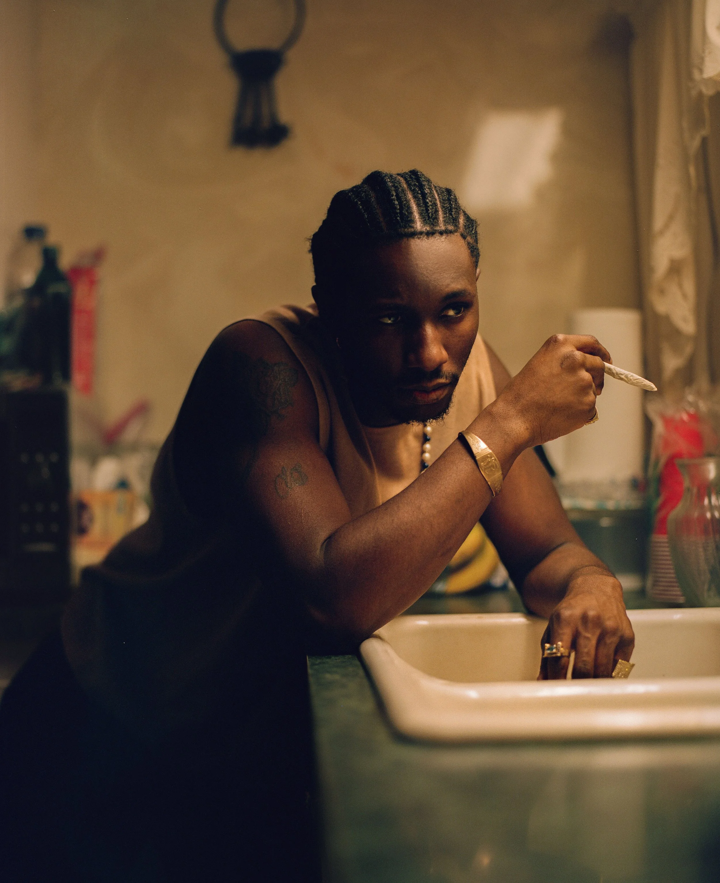 A man with braided hair leaning on a kitchen counter, holding a cigarette, looking pensively. The kitchen is dimly lit with various items in the background.