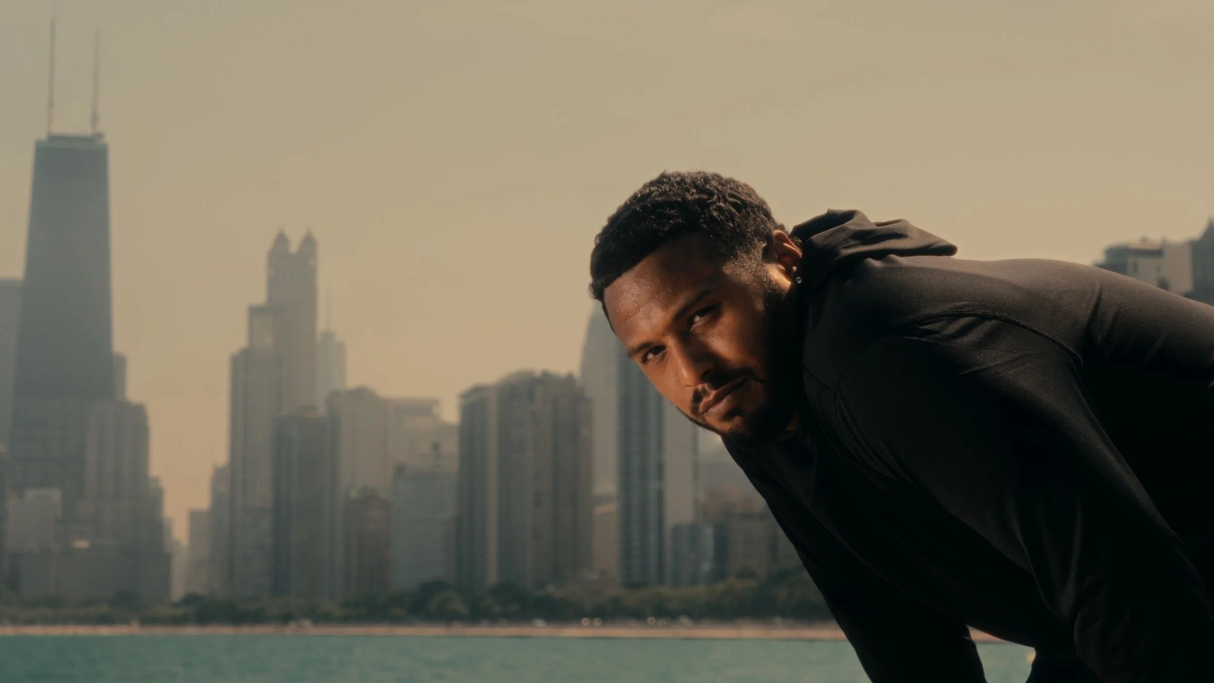 A man in black athletic clothing leaning forward outdoors with a city skyline in the background.