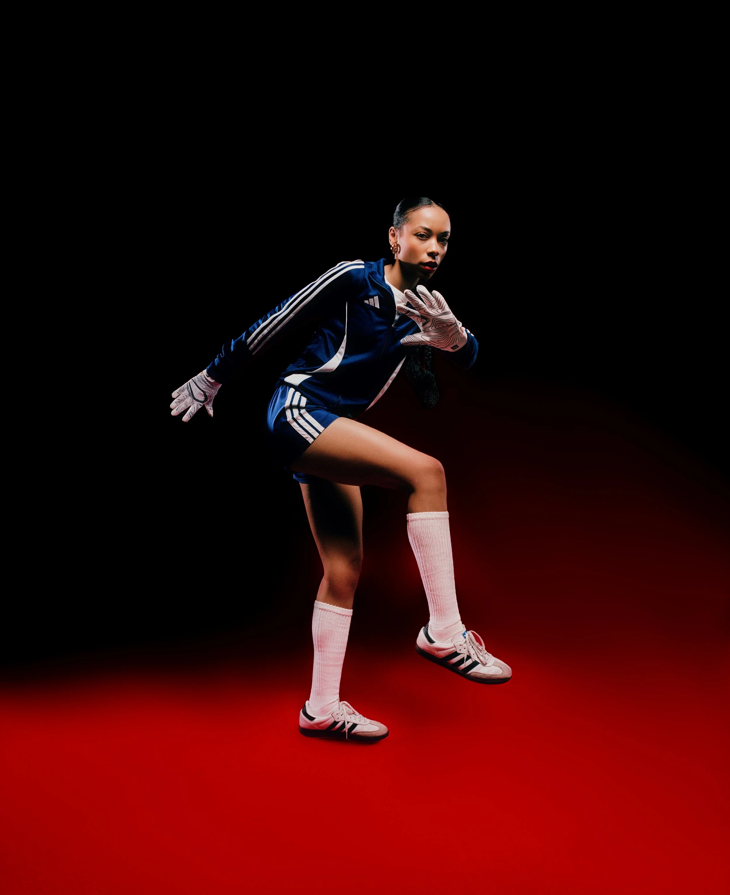 Female soccer player in athletic gear, crouching in a controlled stance against a black background with red floor.