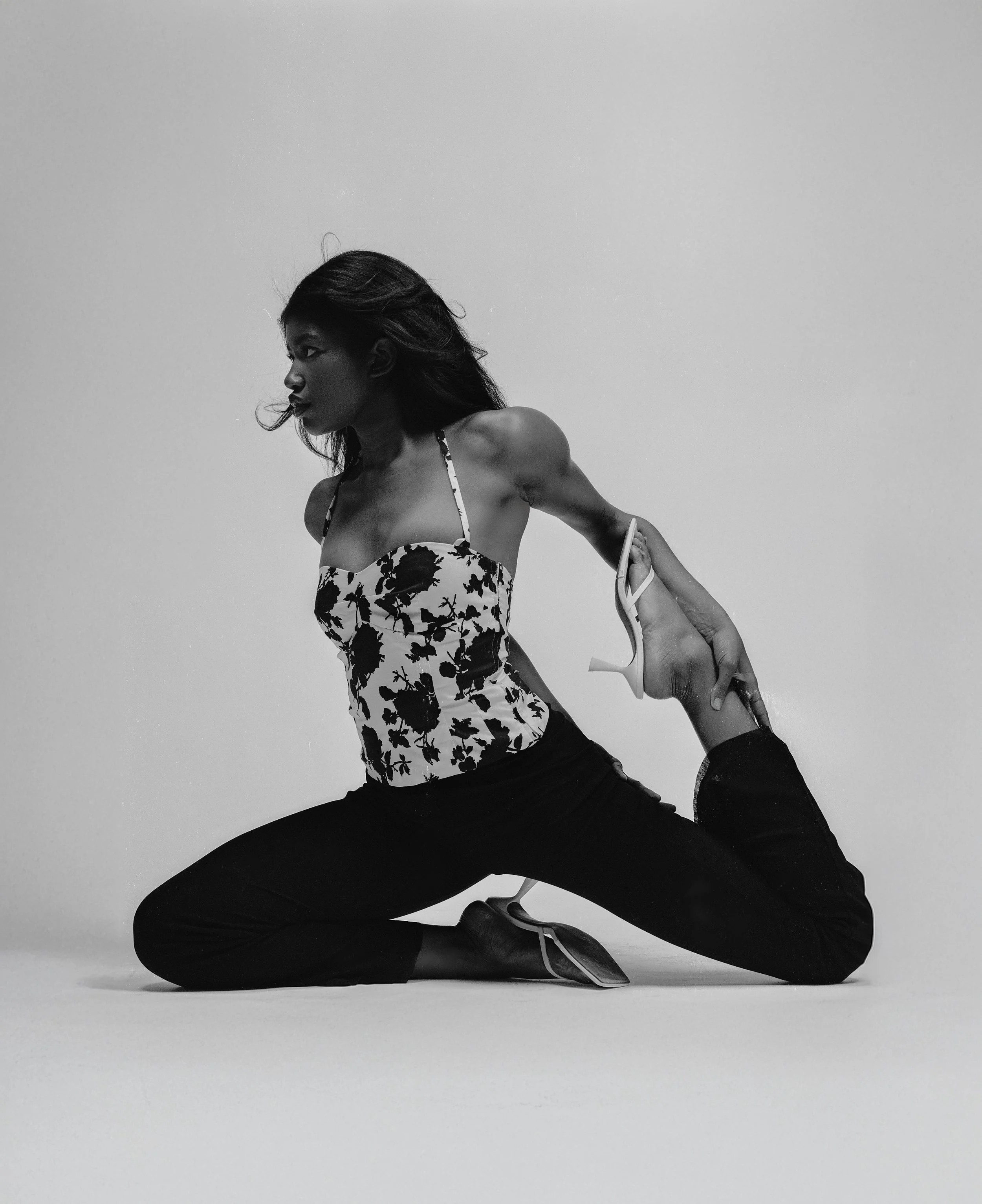 A woman in a floral top and black pants performing a yoga pose, holding her foot behind her head, in a studio with a plain background.