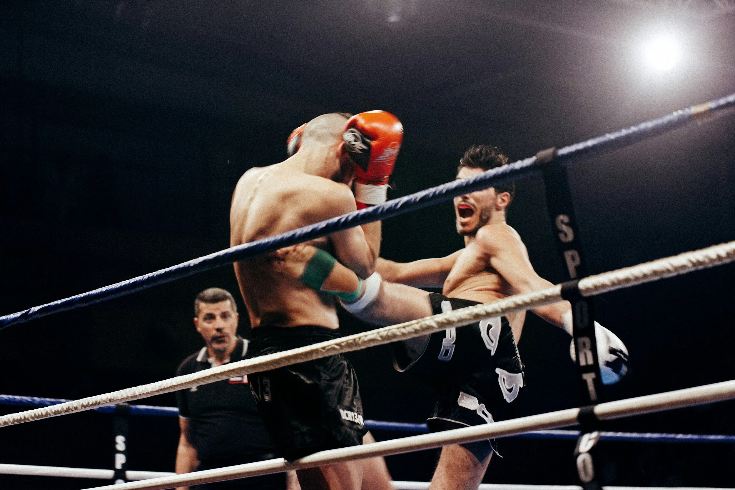 Two male fighters engaged in a kickboxing match; one delivers a kick to the other's midsection, while the other covers his face with gloves; referee observing in the background.