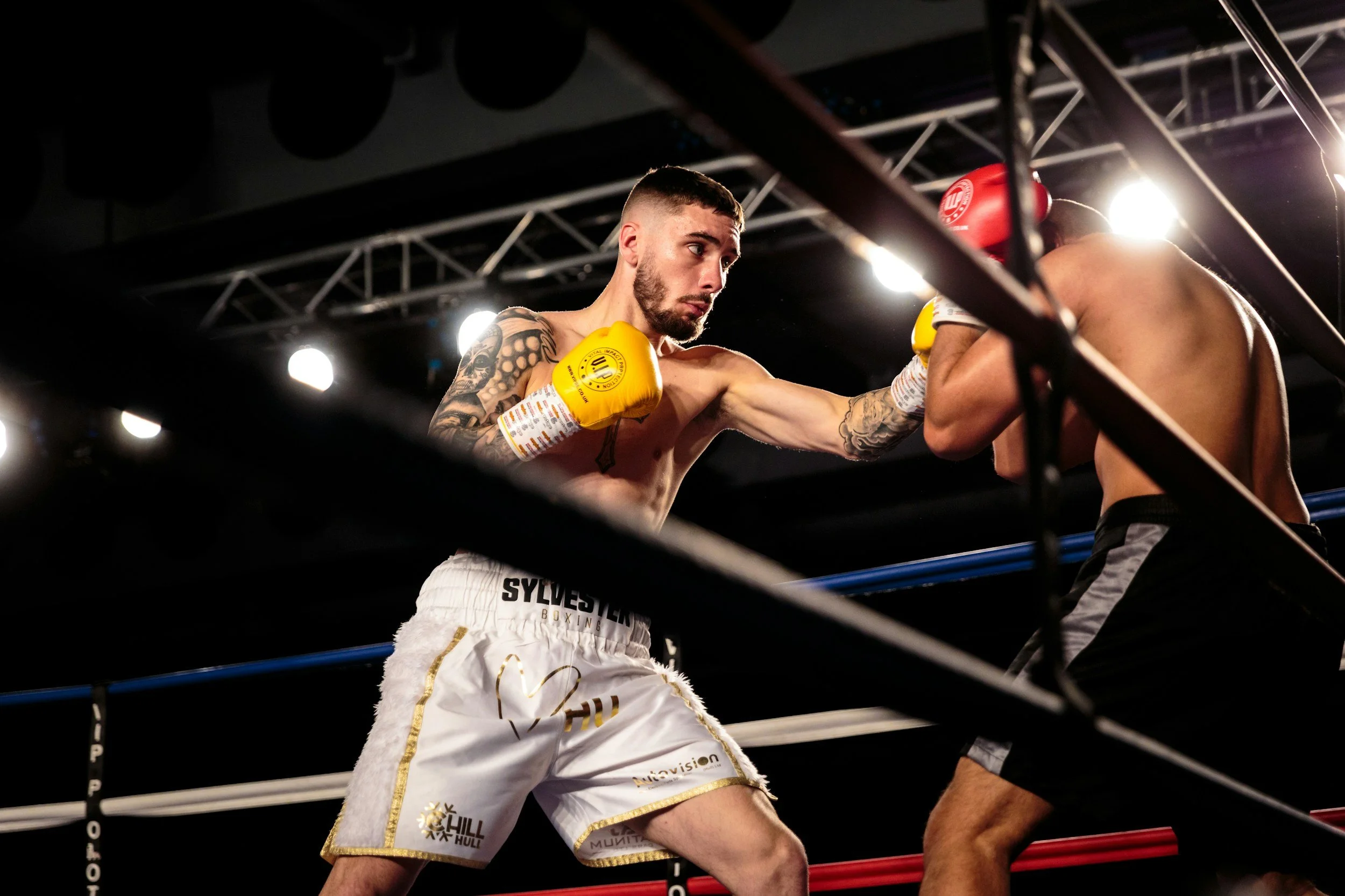 Two shirtless male boxers in a boxing ring; one boxer is throwing a punch while the other is defending. The boxer with yellow gloves has tattoos on his left arm and shorts with various logos. The scene is lit with bright overhead lights.