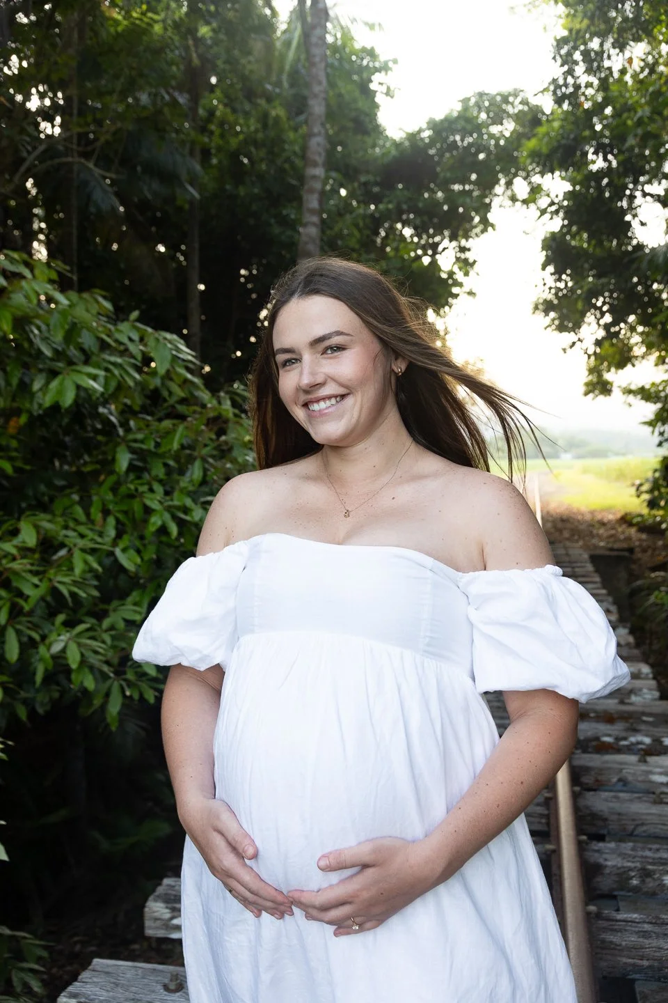 A pregnant woman smiling and holding her belly with both hands, standing outdoors on a wooden bridge surrounded by green foliage and trees during the daytime.