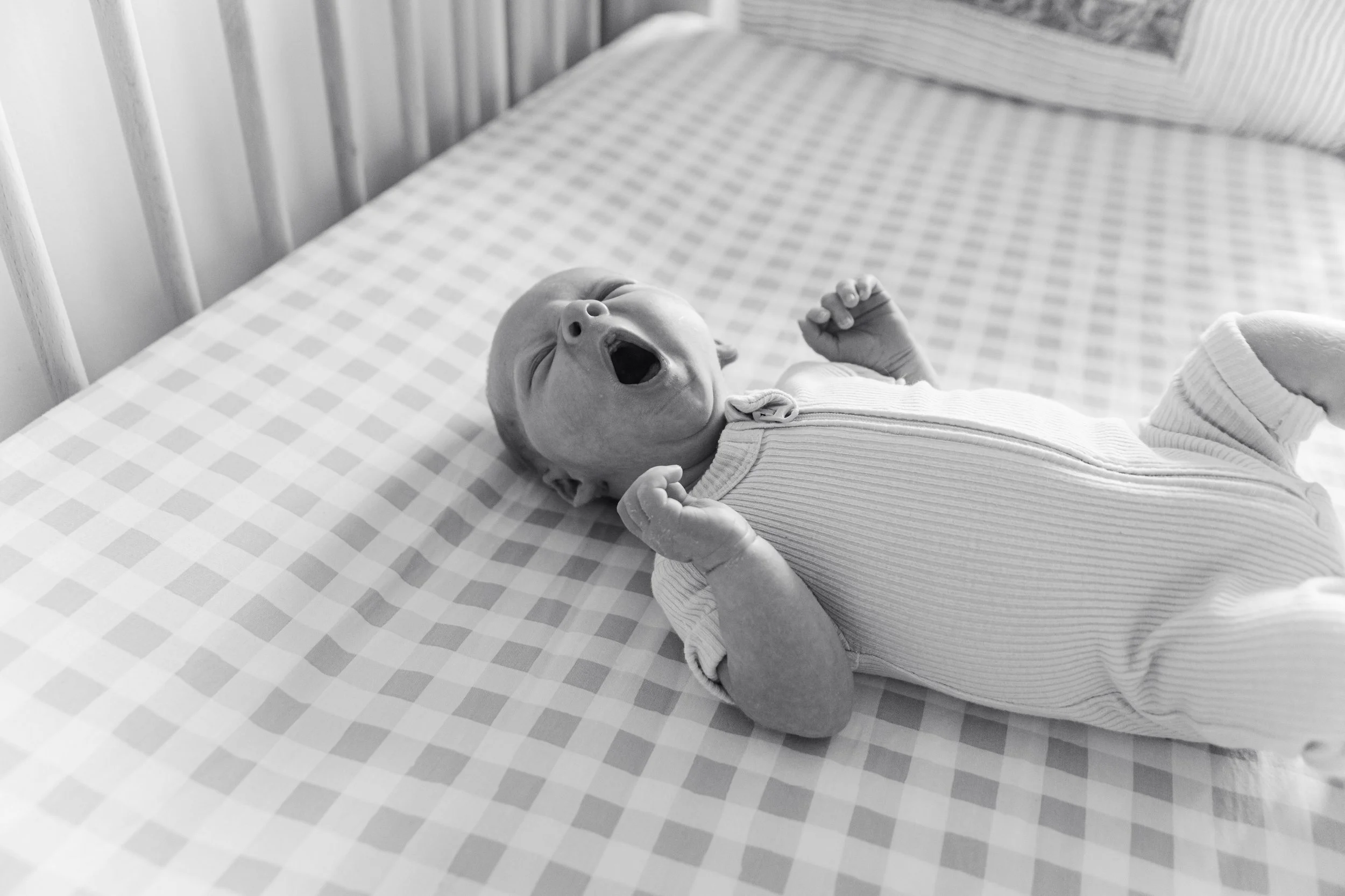 A yawning baby lying on a checkered bed in a crib.