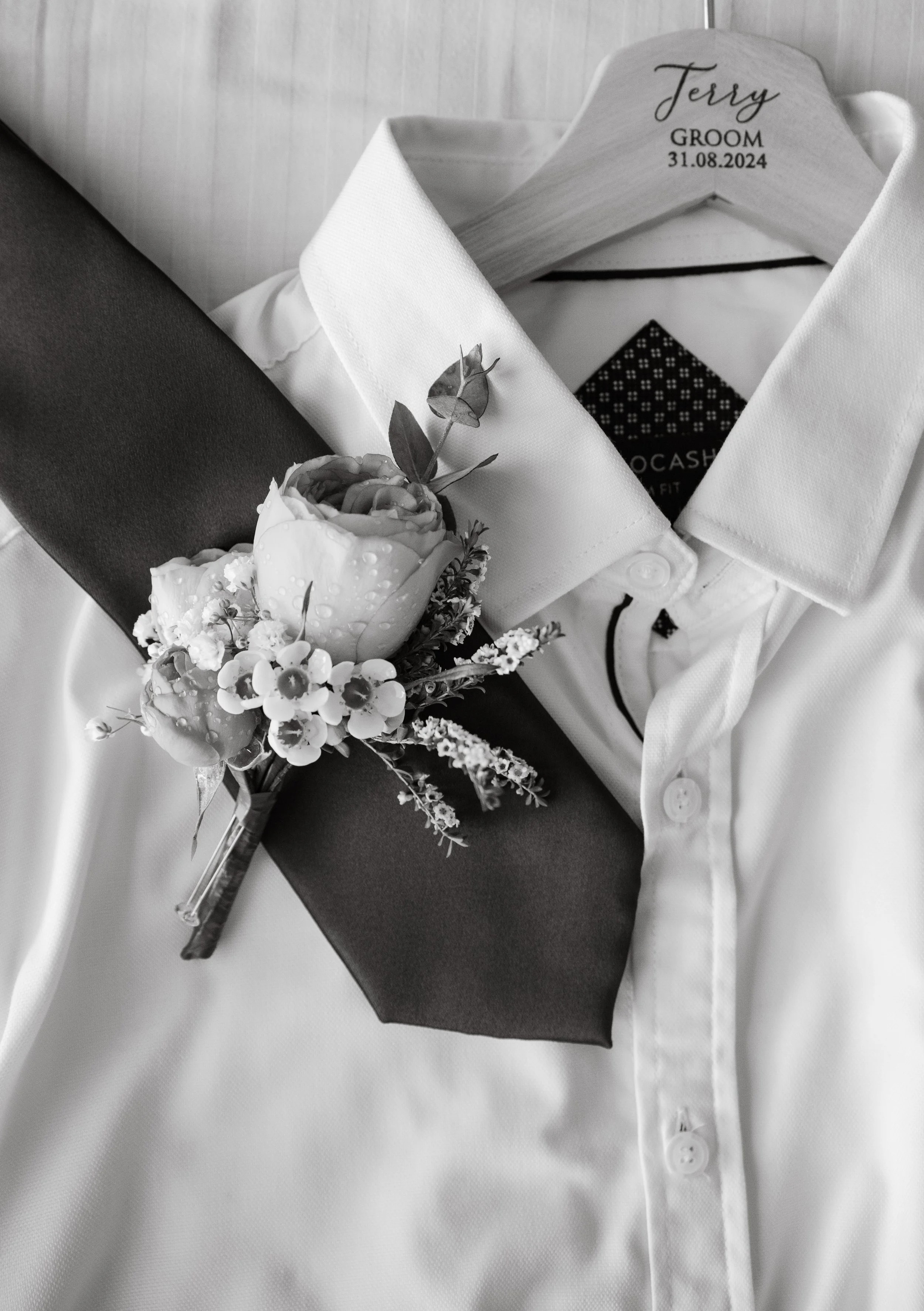 A white dress shirt with a boutonniere resting on the collar and a hanger with the text "Terry Groom 31.08.2024" embroidered, indicating wedding details.