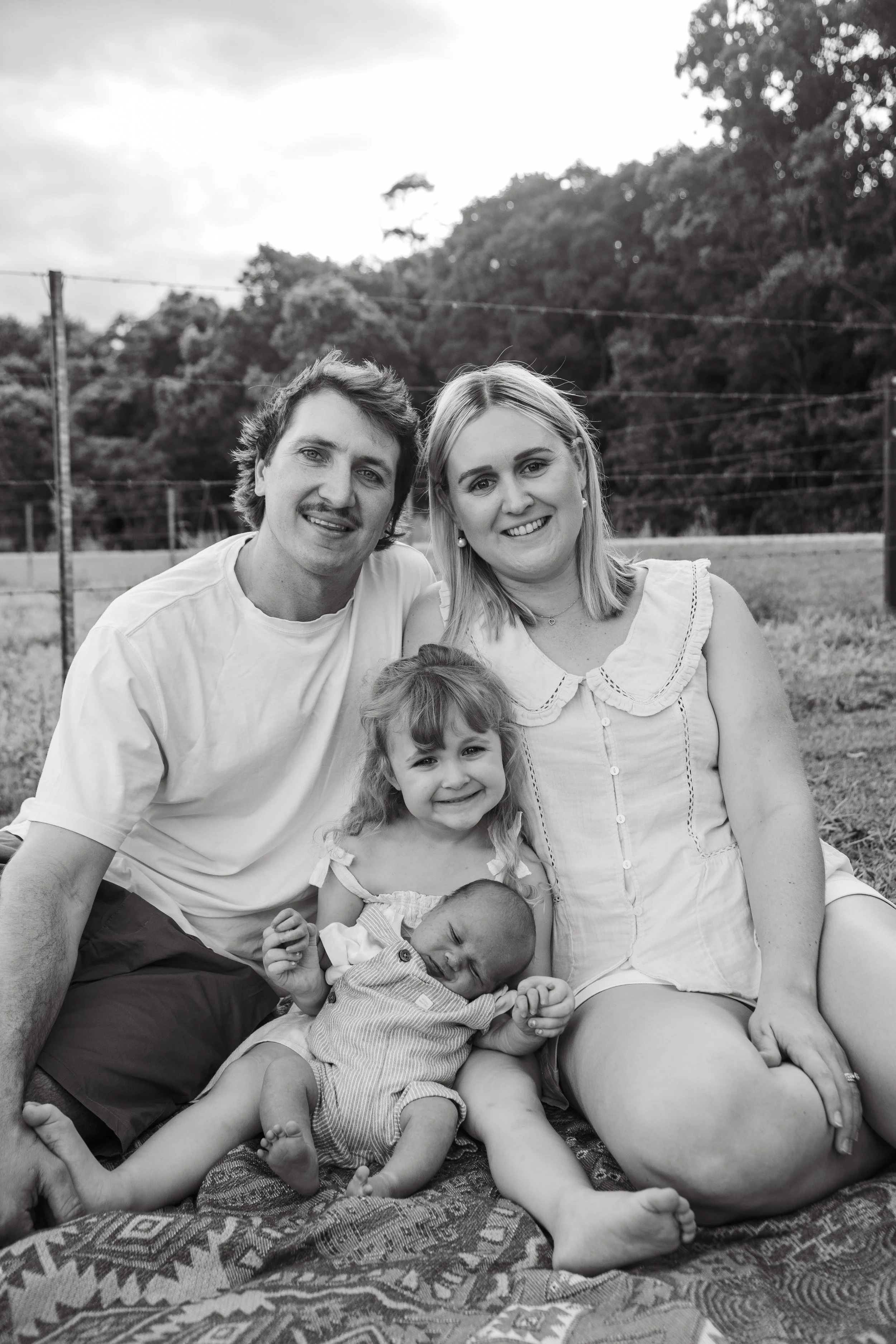 Black and white photo of a family of four, including a father, mother, young girl, and baby, sitting on a blanket outdoors with trees and a cloudy sky in the background.