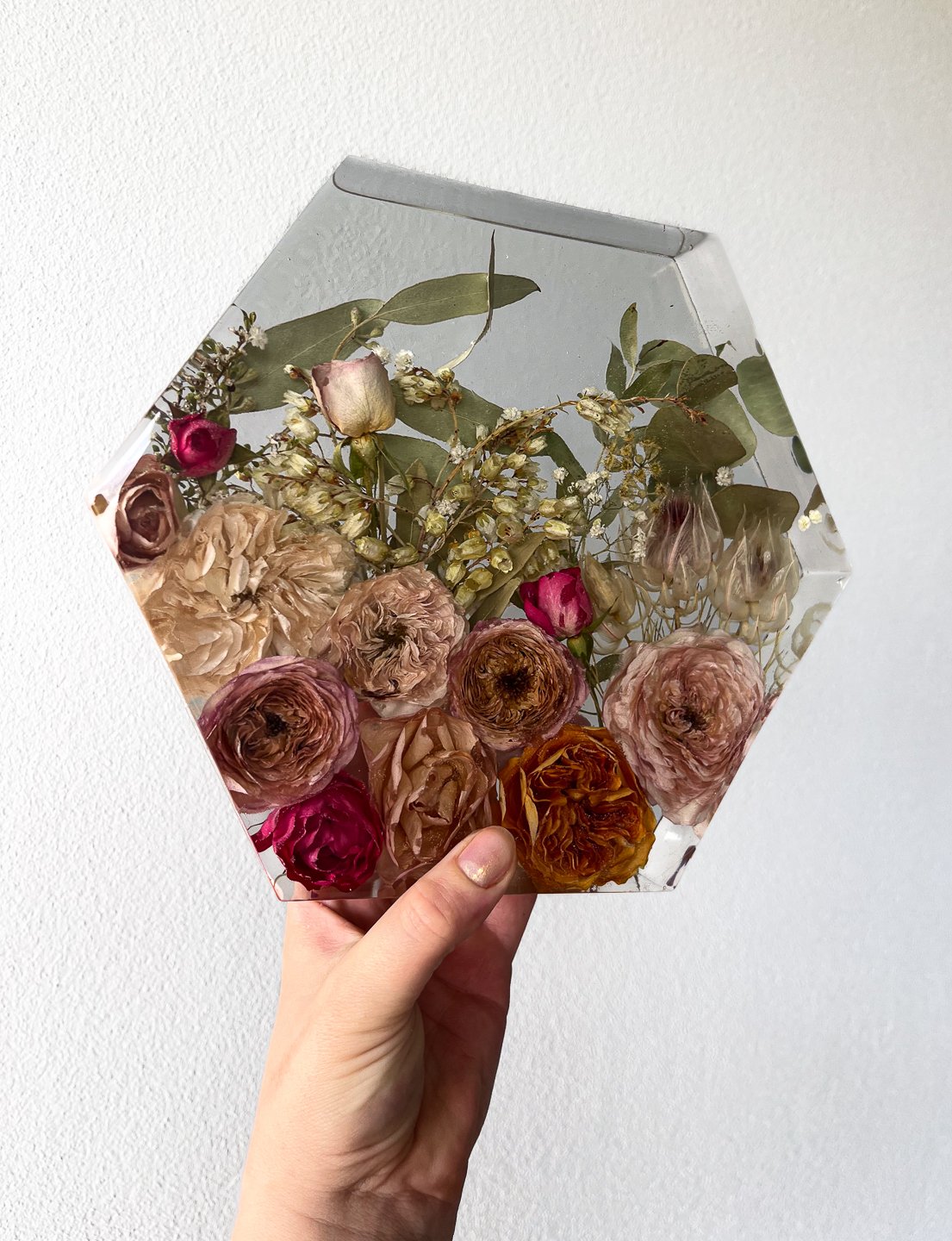 Hand holding a hexagonal glass container with a floral arrangement of preserved dried roses and greenery inside.