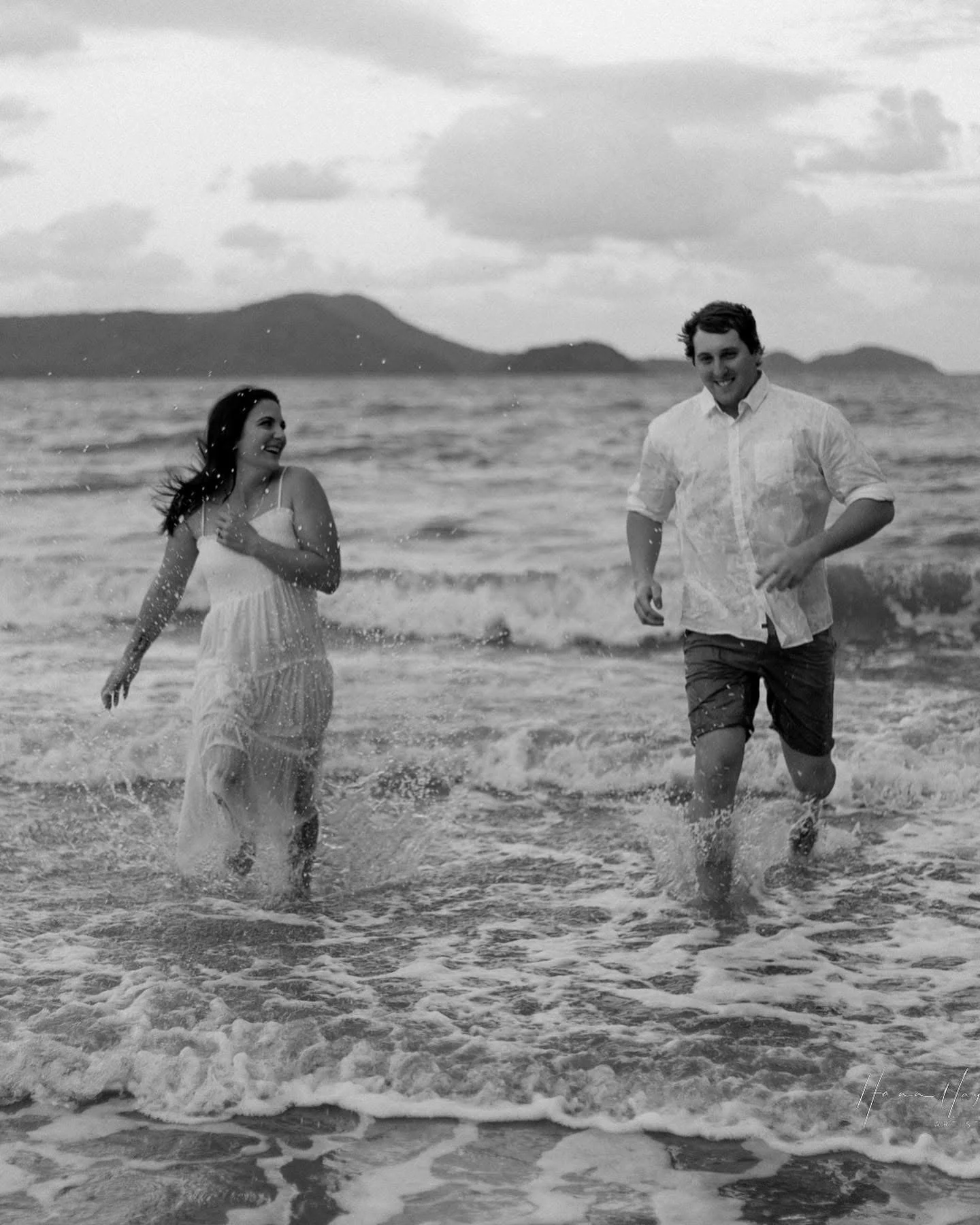 A man and woman running through the ocean waves on a beach, smiling and enjoying themselves, with mountains and clouds in the background.