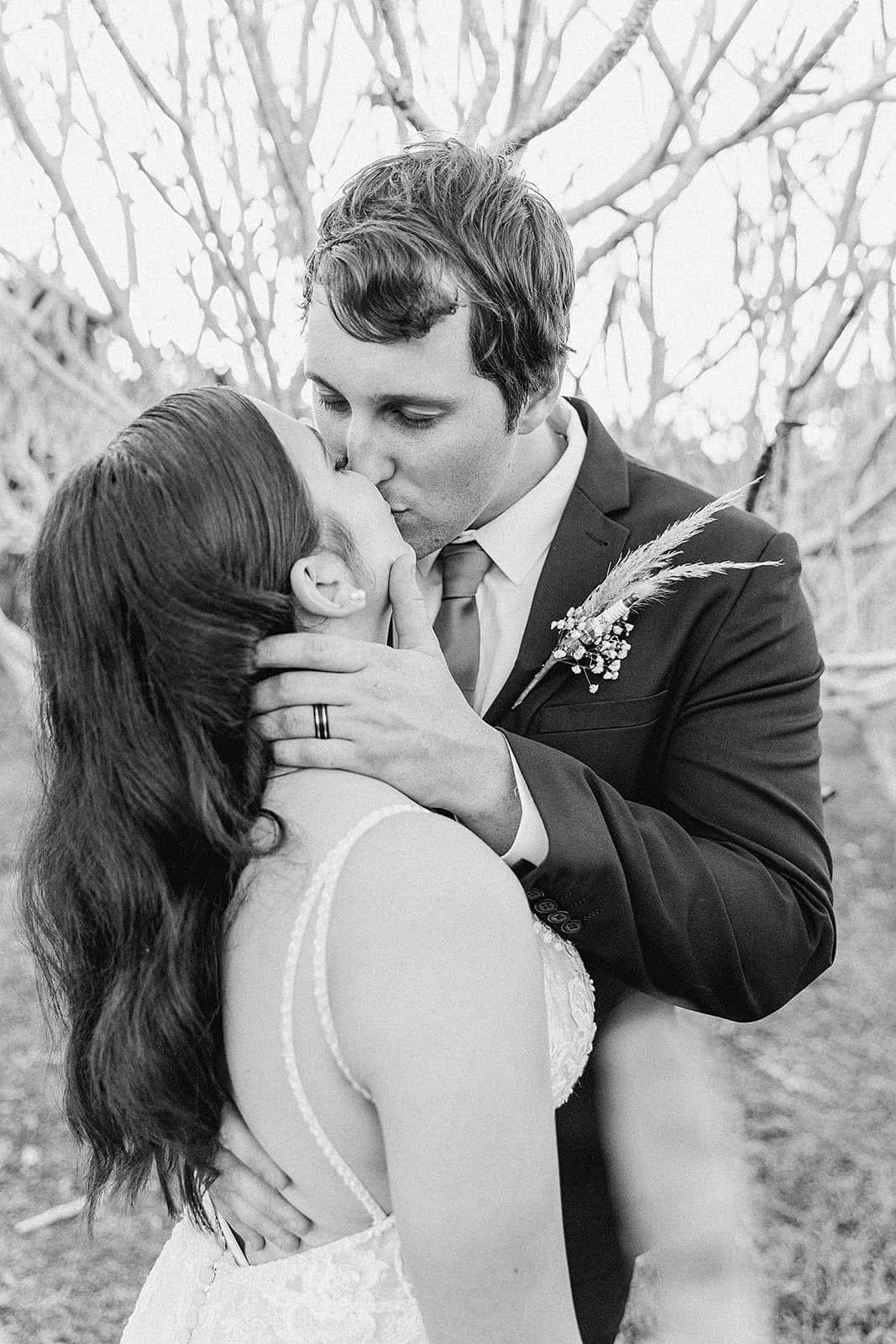 A black and white photo of a couple sharing a kiss, outdoors with bare tree branches in the background. The man is dressed in a suit with a boutonniere, and the woman is wearing a white dress with thin straps.