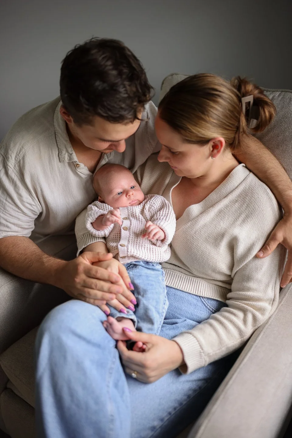 A family of three sitting on a beige sofa, with the father and mother looking at their baby who is sitting on the mother's lap. The baby is wearing a hand-knitted sweater and has wide eyes.