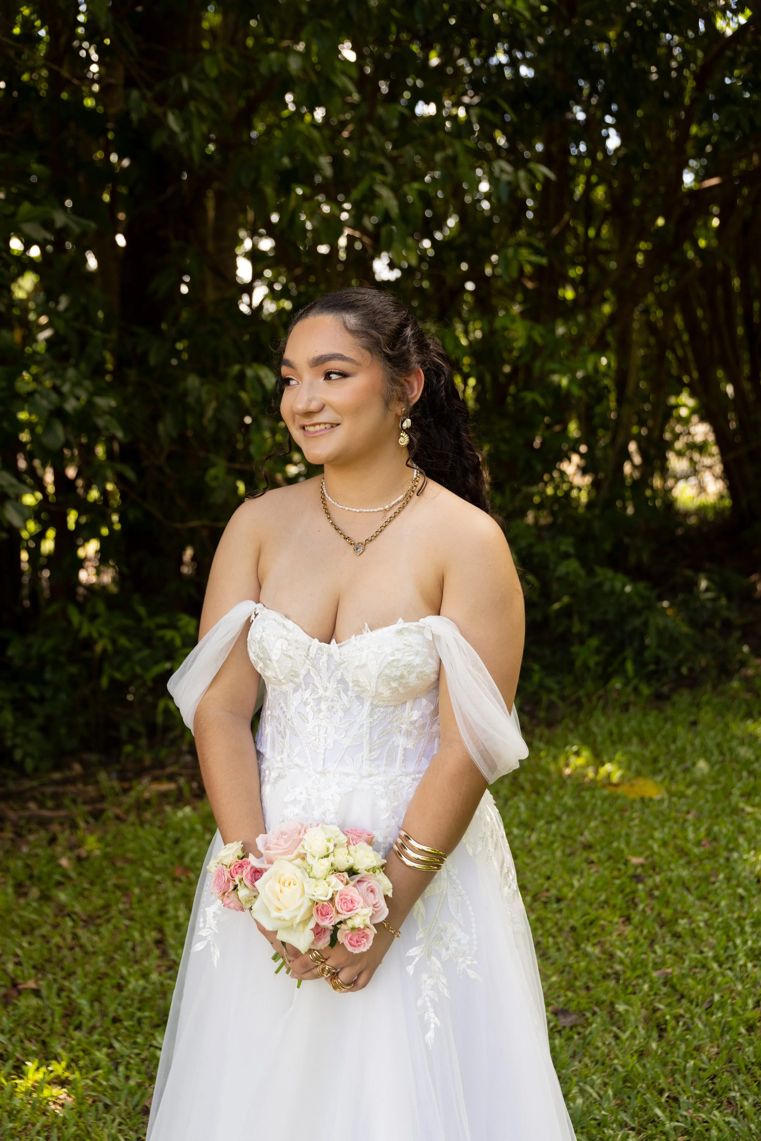 A young woman in a white wedding dress holding a bouquet of pink and white roses, standing outdoors with green foliage background.