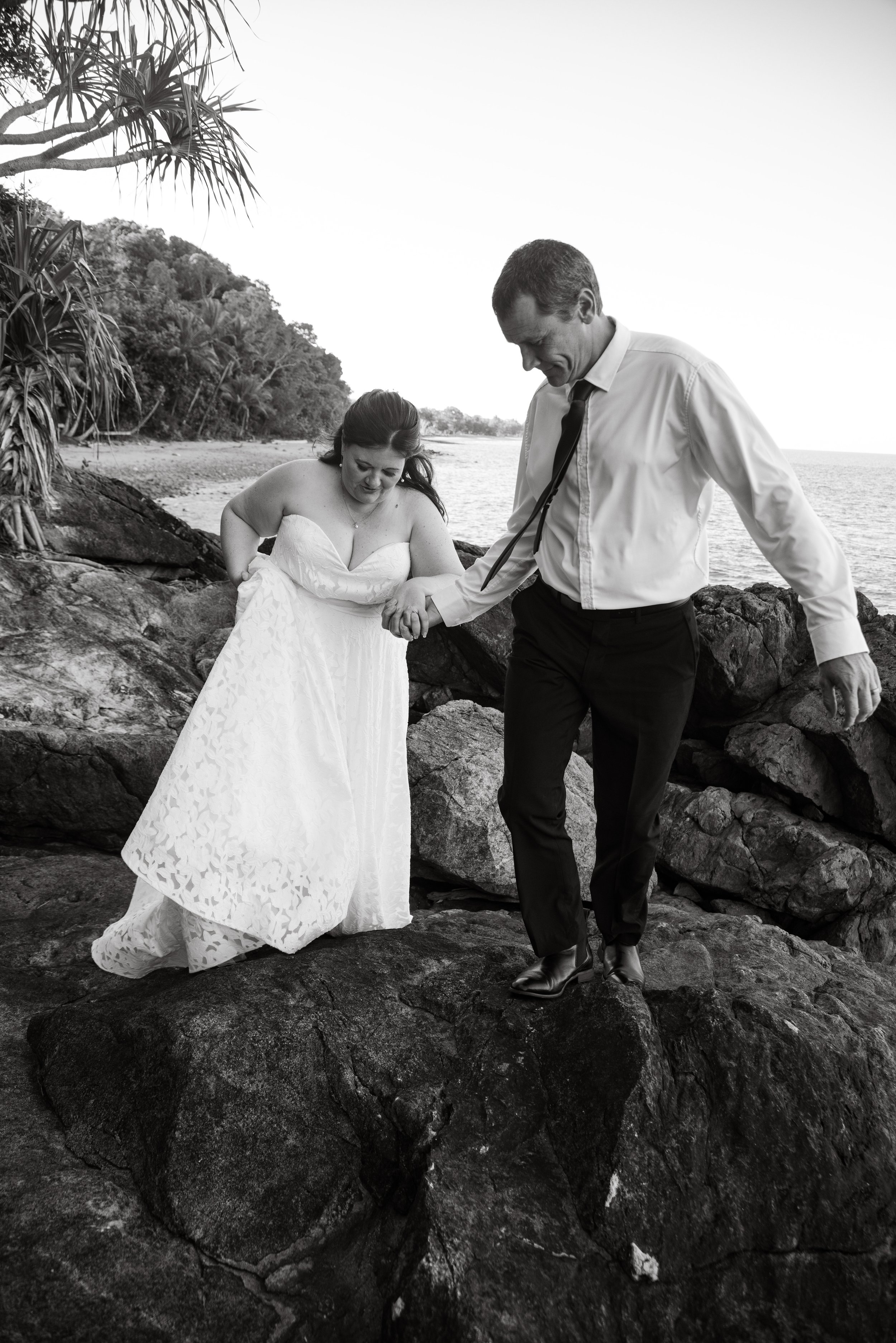 A bride in a white dress and a groom in a shirt and tie walking together on rocks by the beach, holding hands.