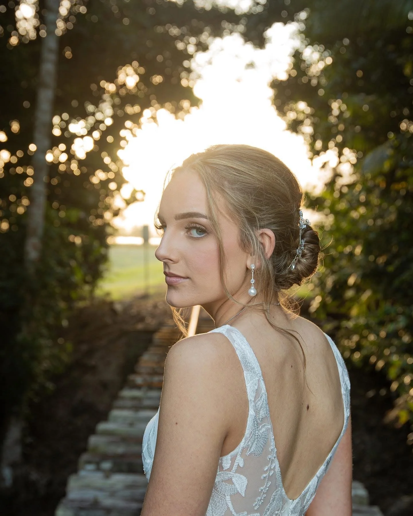 A woman with blonde hair in an updo and pearl earrings looking over her shoulder on a wooden bridge at sunset.