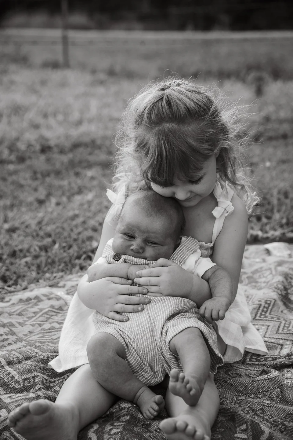 A young girl hugging a baby boy outdoors on a patterned blanket.