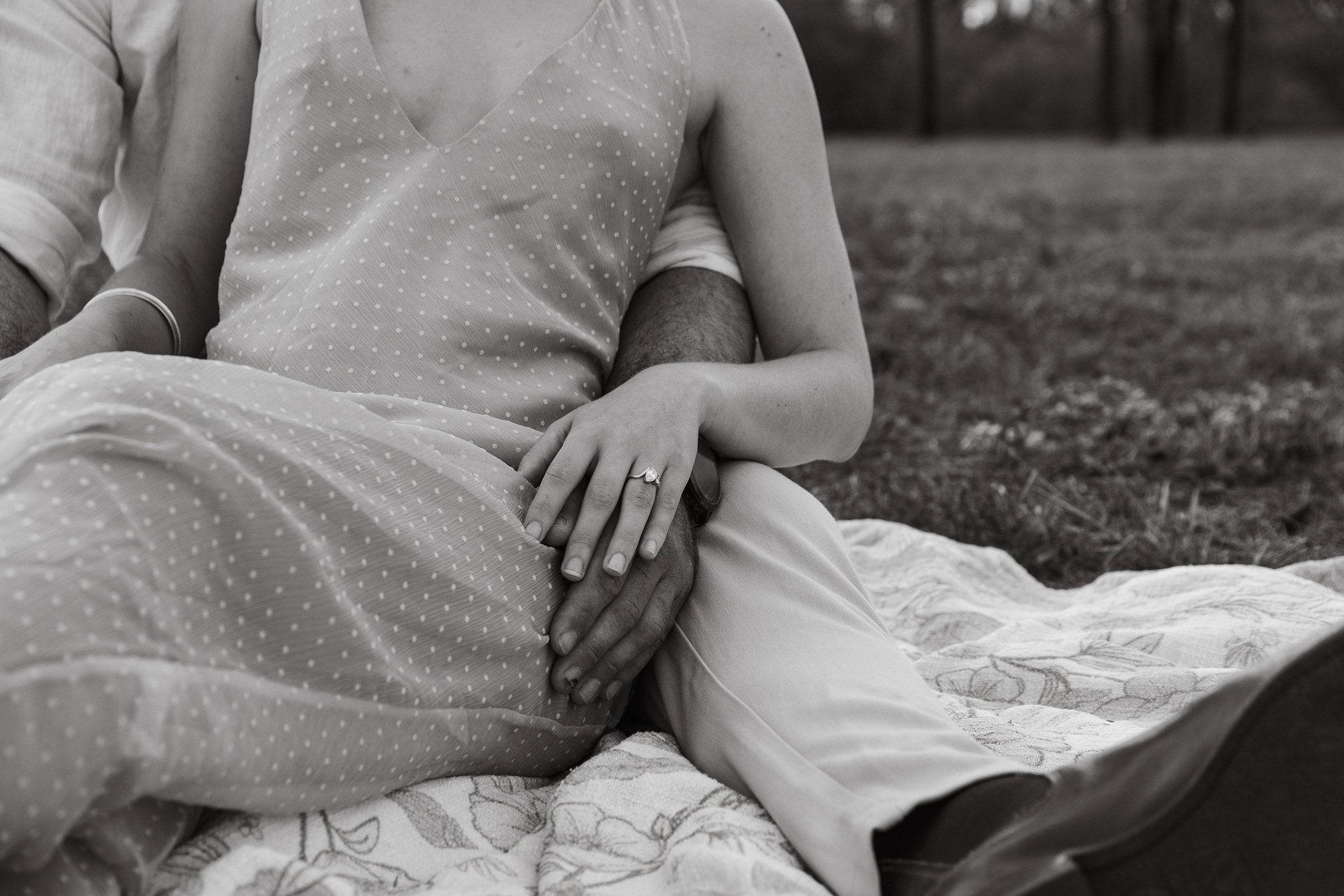 Close-up of couple sitting outdoors, holding hands on lap, with a ring on the woman's finger, black-and-white photo.