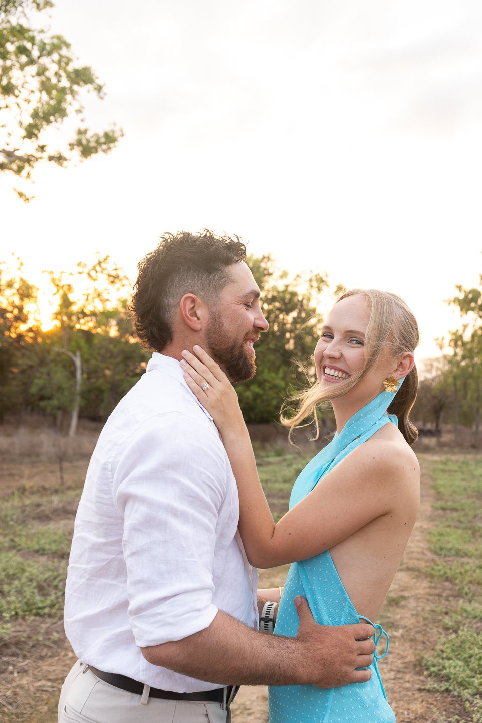 A smiling couple embracing outdoors during sunset, the woman holding the man's face lovingly, in a natural setting with trees in the background.