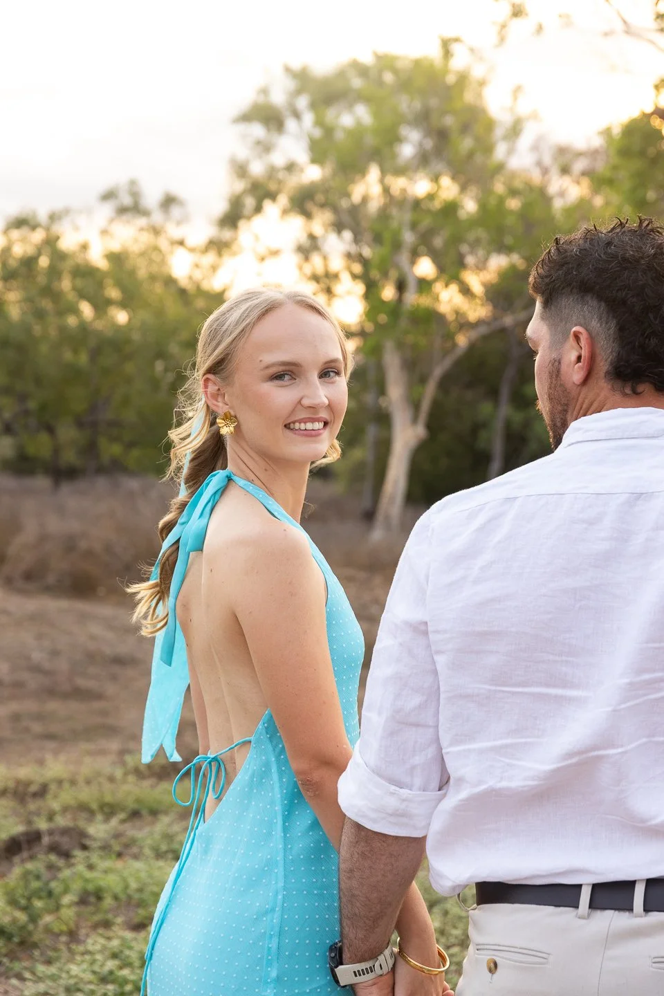 A woman with blonde hair in a loose braid, wearing a blue sleeveless dress with polka dots and gold earrings, smiles at a man in a white shirt in an outdoor setting with trees and a sunset sky in the background.