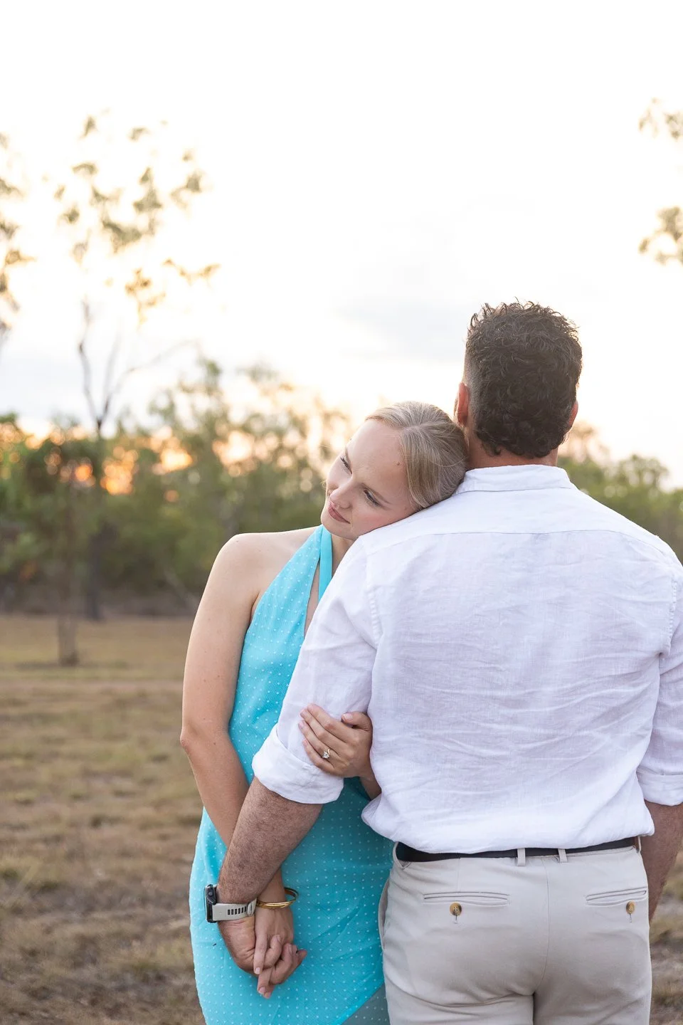 A woman hugging a man from behind outdoors during sunset, with trees in the background.
