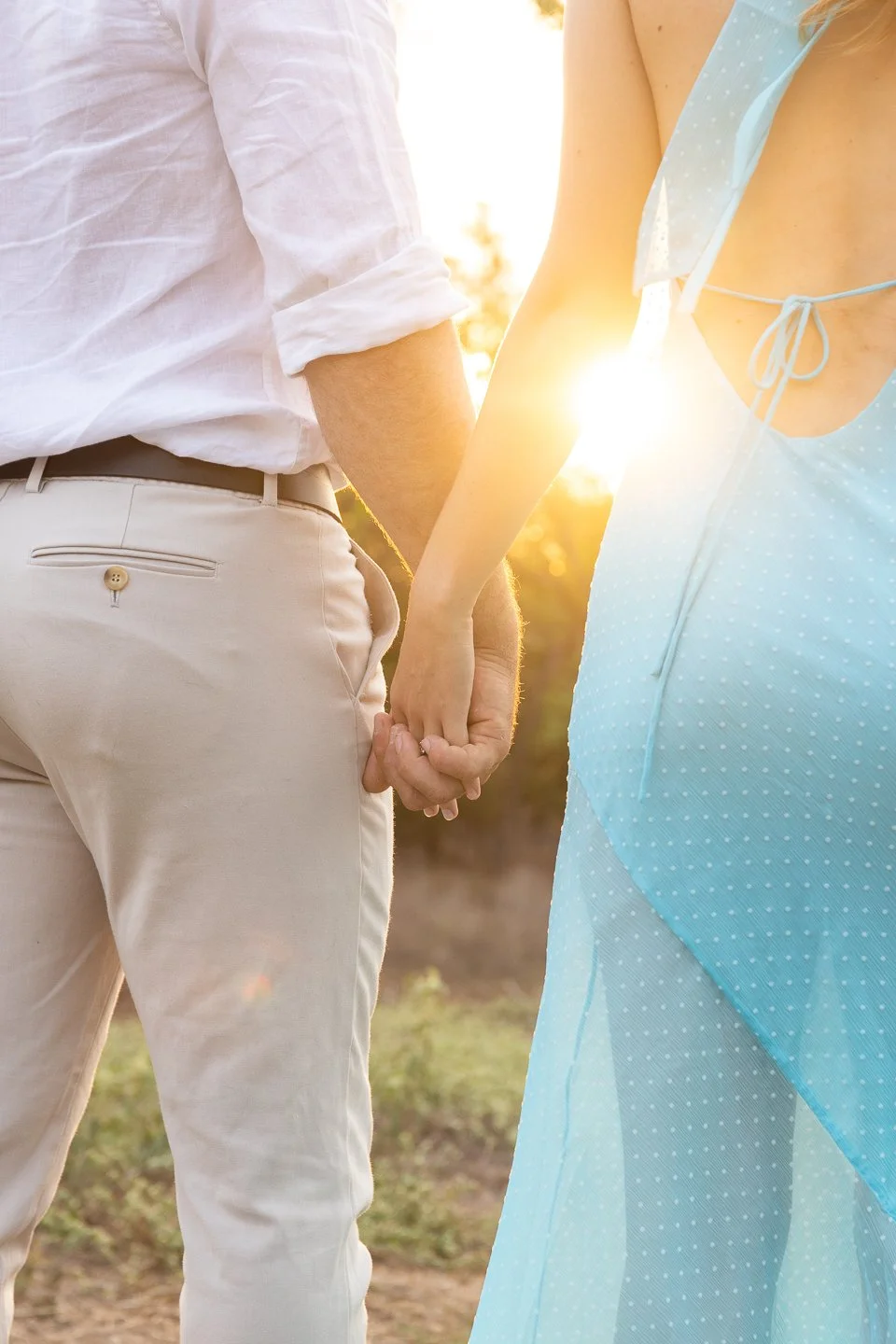 A couple holding hands outdoors at sunset, with the person's face not visible, emphasizing their hands and intertwined fingers.