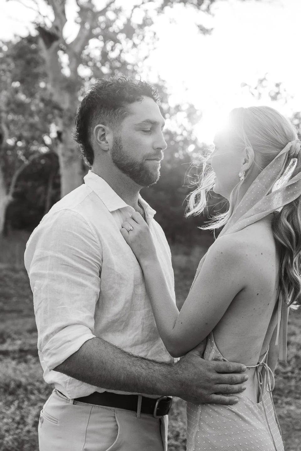 A black and white photo of a couple holding each other closely outdoors, with trees in the background and sunlight shining behind them.