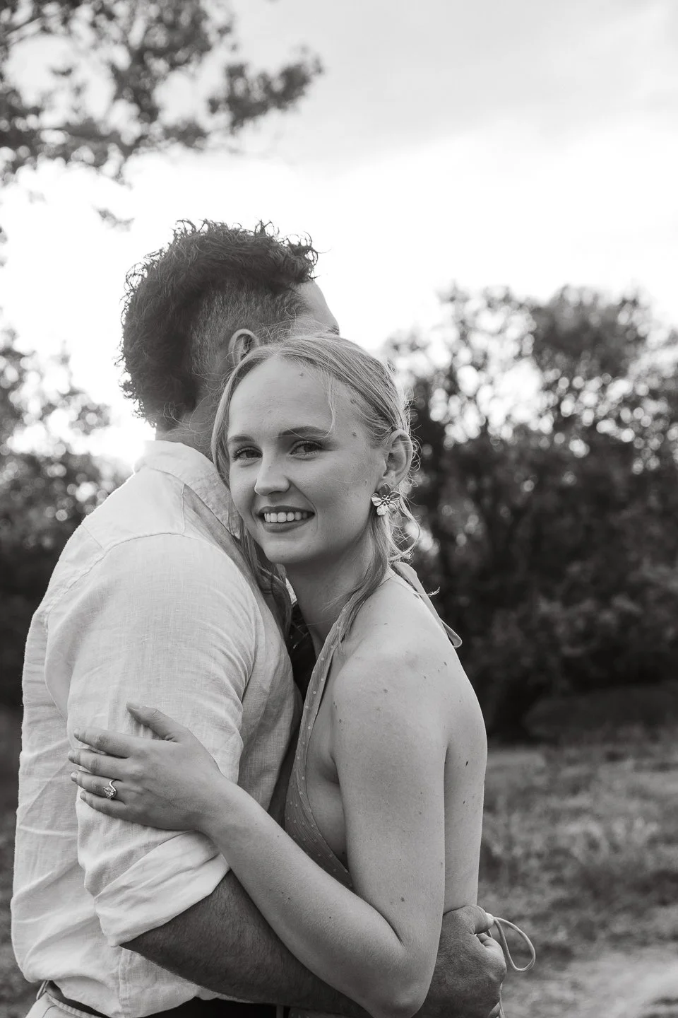 Black and white photo of a smiling woman with earrings being embraced by a man, outdoors with trees in the background.
