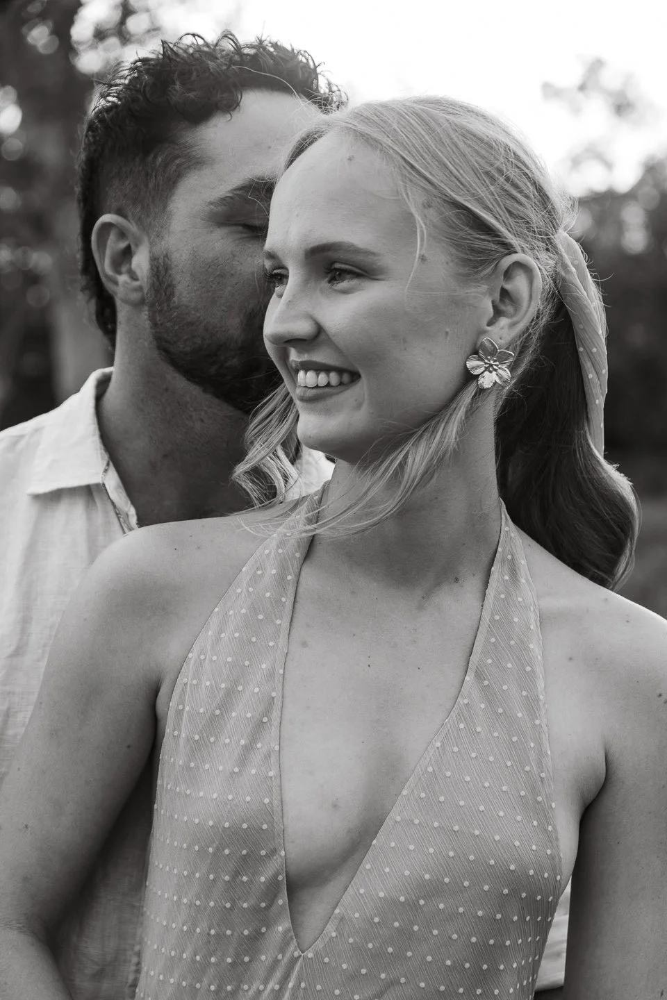 A black-and-white photo of a smiling blonde woman with flowers earrings and a deep V polka-dot dress, with a man whispering into her ear, outdoor setting.