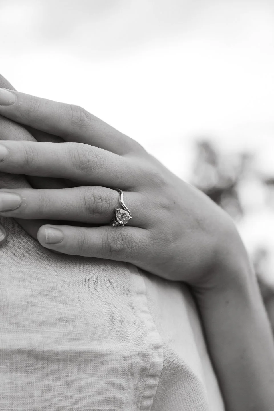 Close-up of a woman wearing a diamond engagement ring on her ring finger, resting her hand on her shoulder with a blurred background.