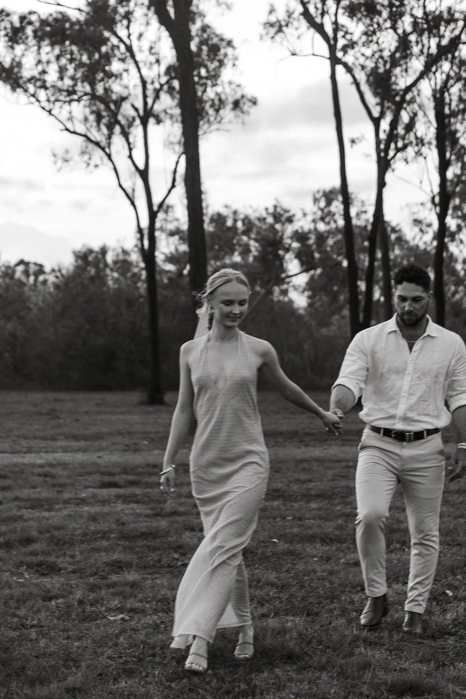 A black and white photo of a man and woman walking on grass in a park or forested area, holding hands, with tall trees in the background.