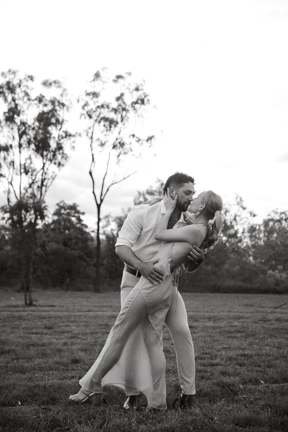 A black-and-white photo of a couple kissing and embracing outdoors in a park with trees in the background.