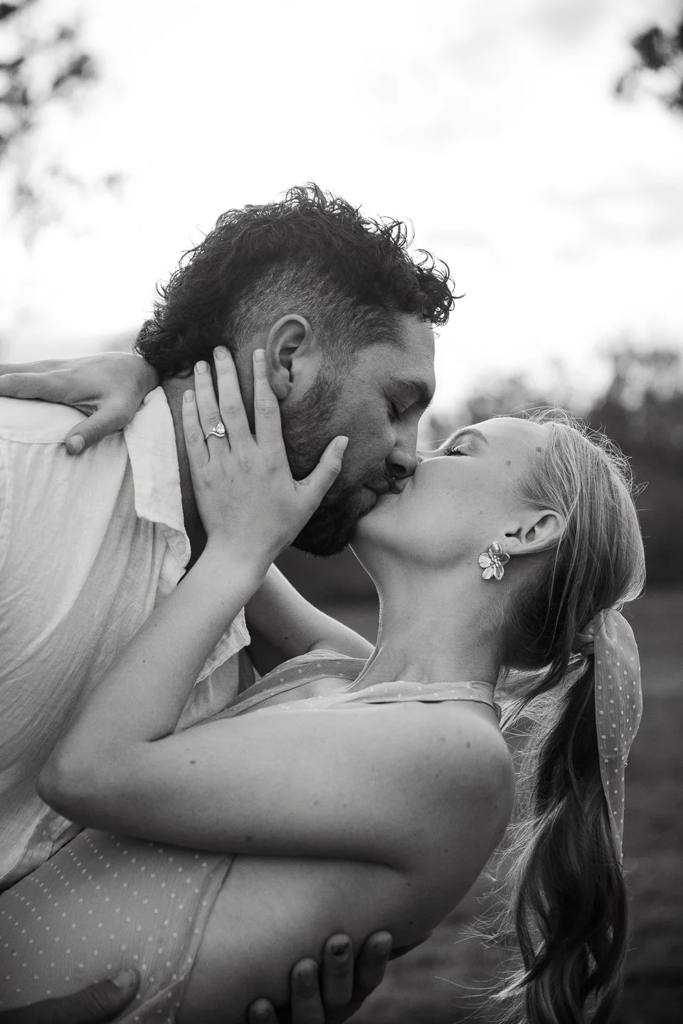 Black and white photo of a man and woman kissing outdoors. The woman holds the man's face with one hand, showing an engagement ring. The woman's side profile is visible, with earrings and a ponytail. The man's face is partially visible, with a beard and curly hair.