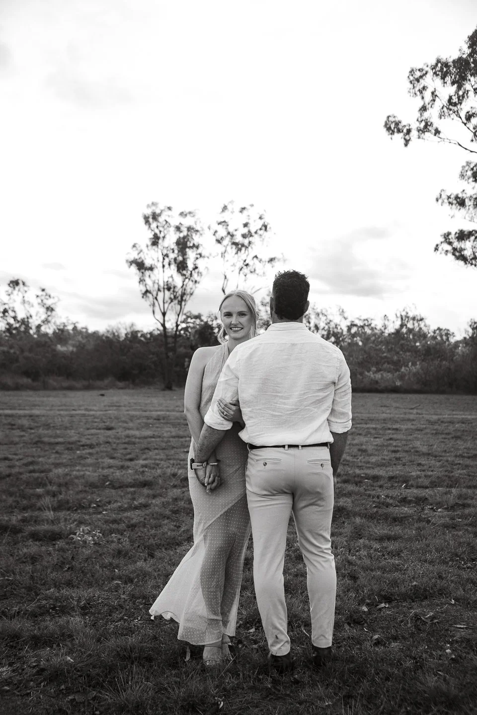 A black and white photo of a smiling woman in a dress holding hands with a man in a light shirt and pants, standing outdoors on a grassy field with trees and a cloudy sky in the background.