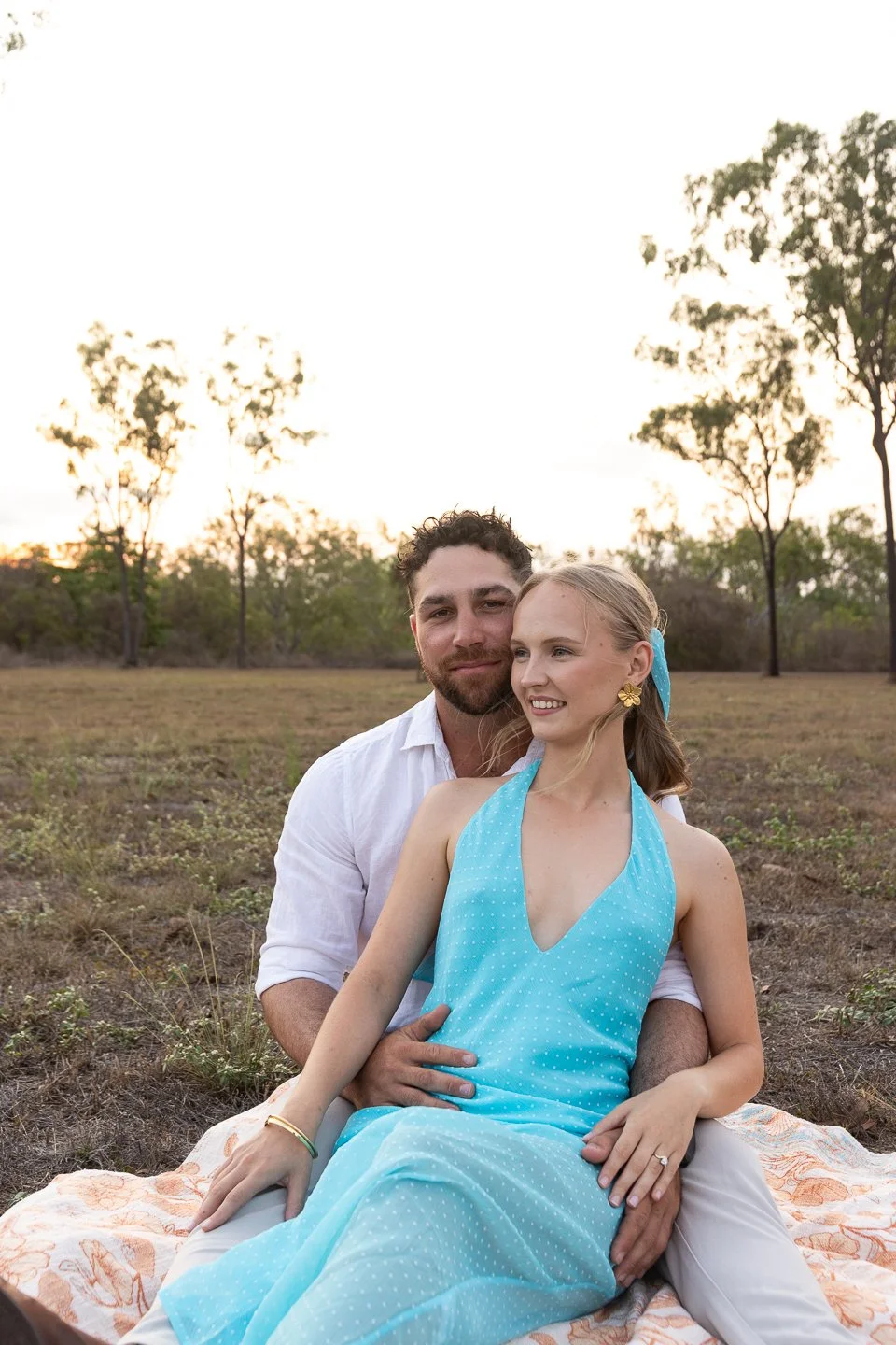 A couple sitting on a blanket outdoors in a field with trees in the background during sunset, enjoying a romantic moment.