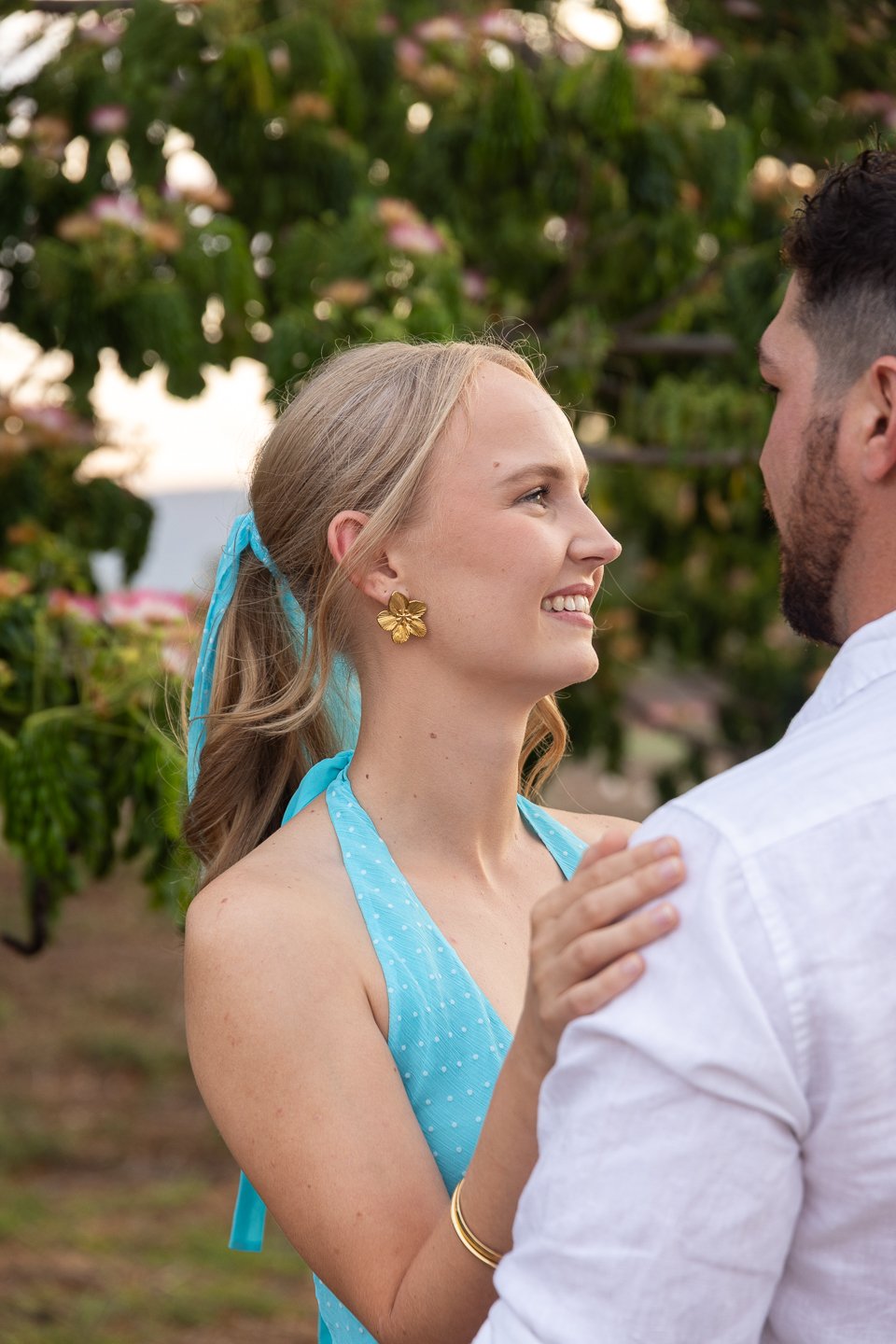 A woman with blonde hair tied back with a blue ribbon, wearing gold flower-shaped earrings and a blue dress with white polka dots, smiles at a man with dark hair and a beard, wearing a white shirt, outdoors among green foliage and pink flowers.