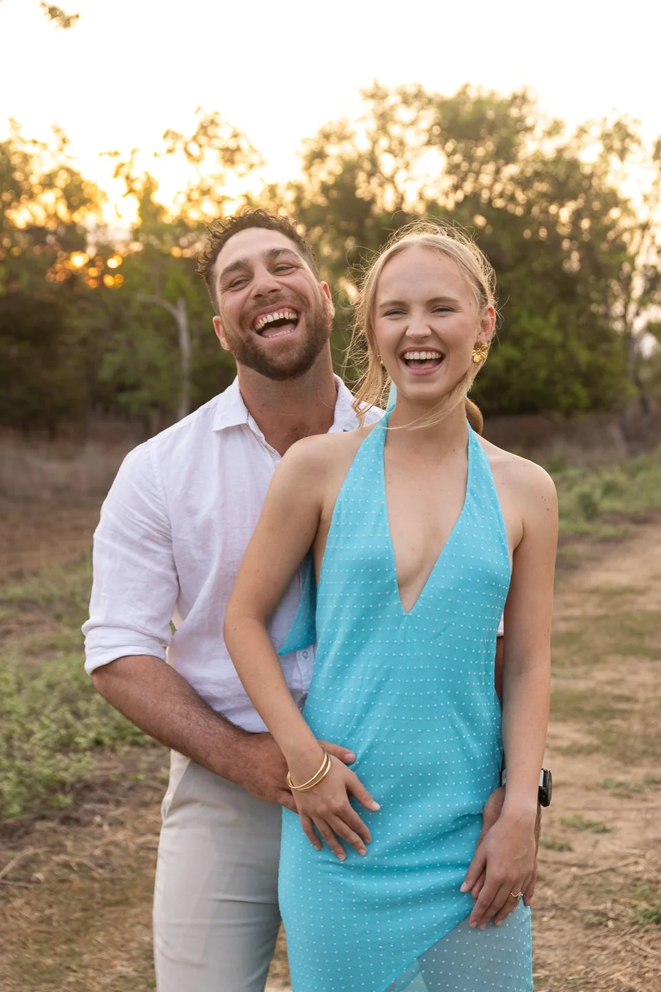 A happy man and woman laughing outdoors at sunset, standing in a field with trees in the background. The woman is wearing a blue halter dress and the man is in a white shirt.