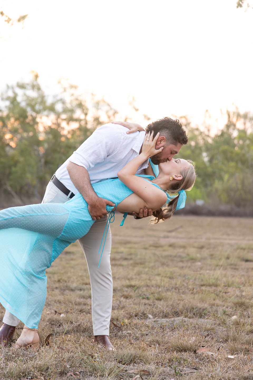 A couple is sharing a romantic kiss outdoors. The woman is being dipped by the man, and they appear happy and in love.
